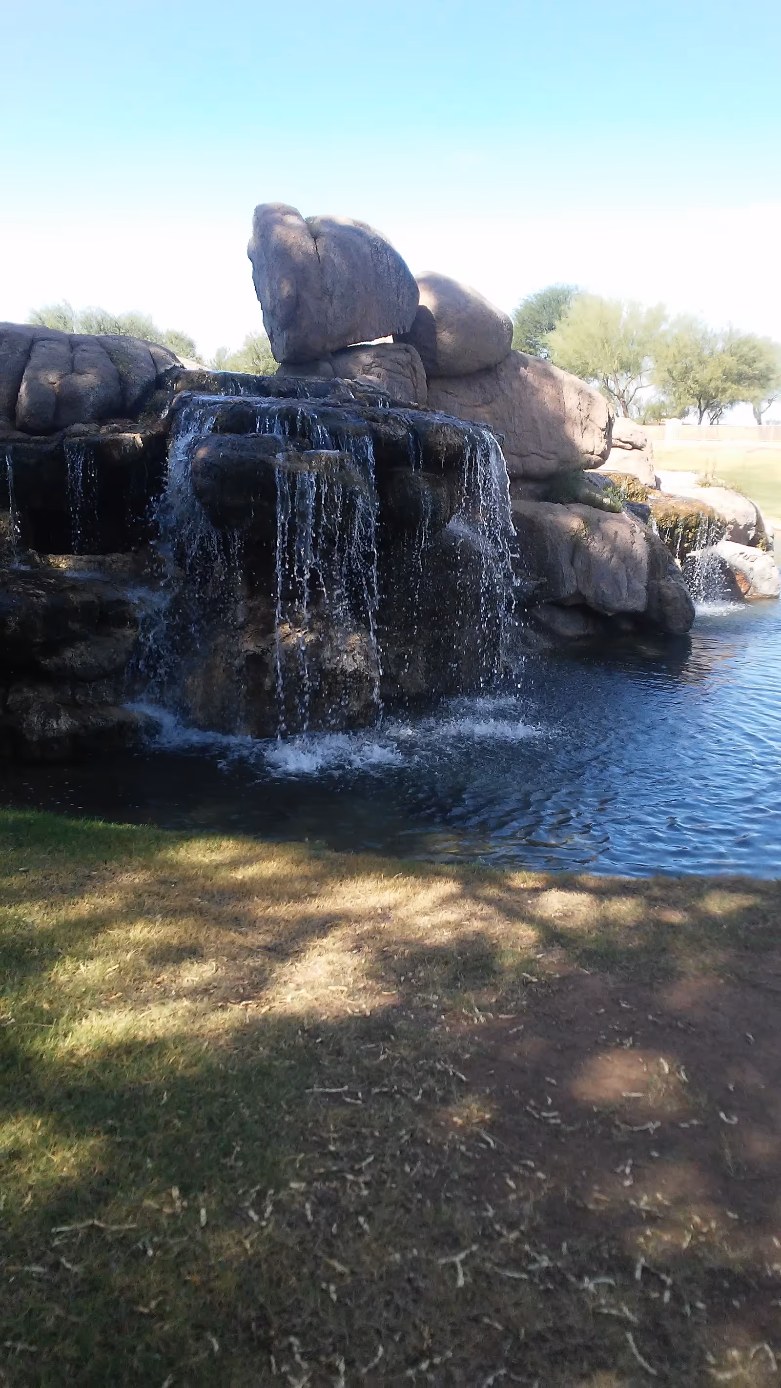 Rock waterfall over a pond with grassy foreground and distant trees under blue sky.