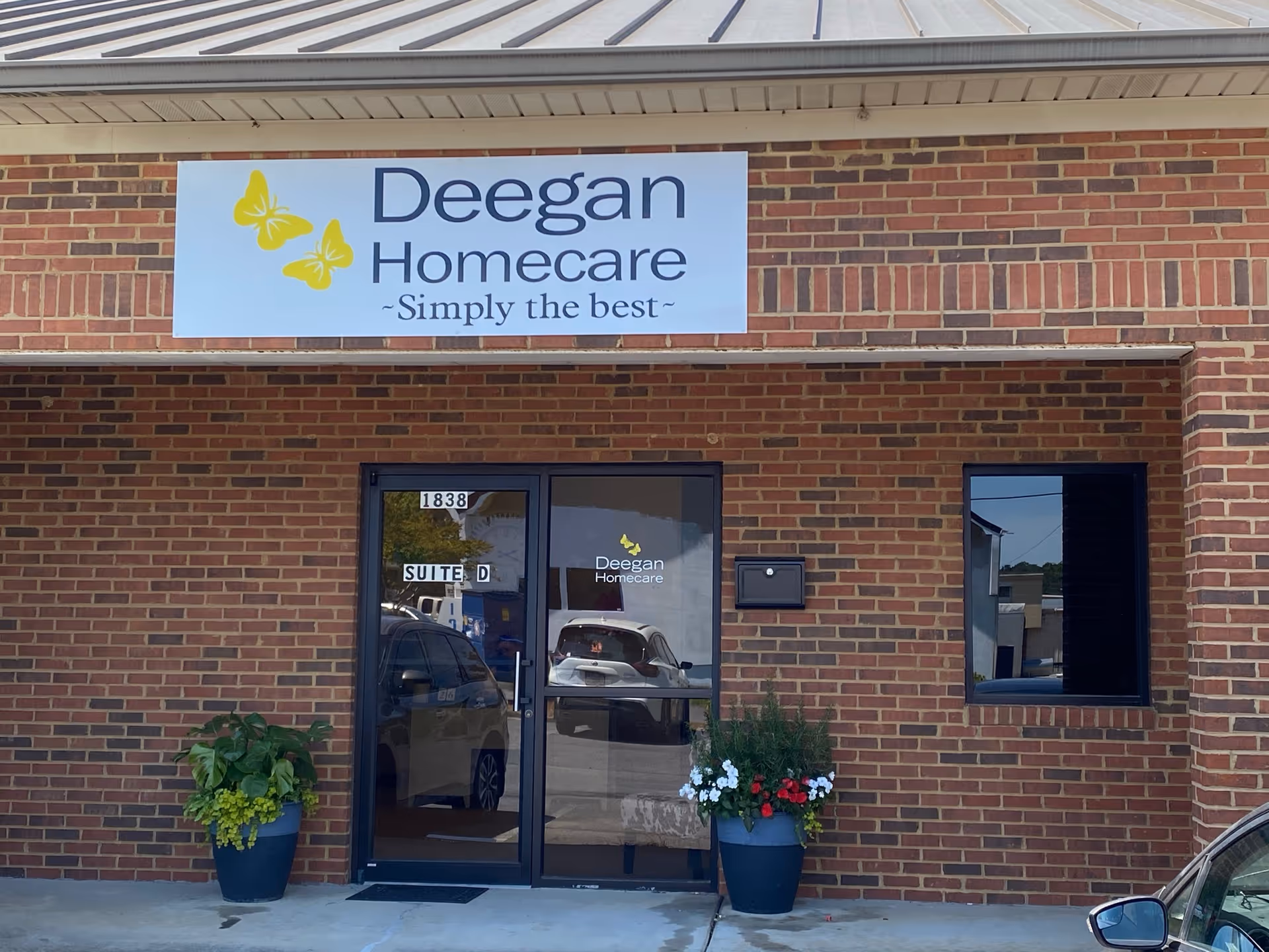 Brick facility entrance with Deegan Homecare sign, glass doors, and potted plants.