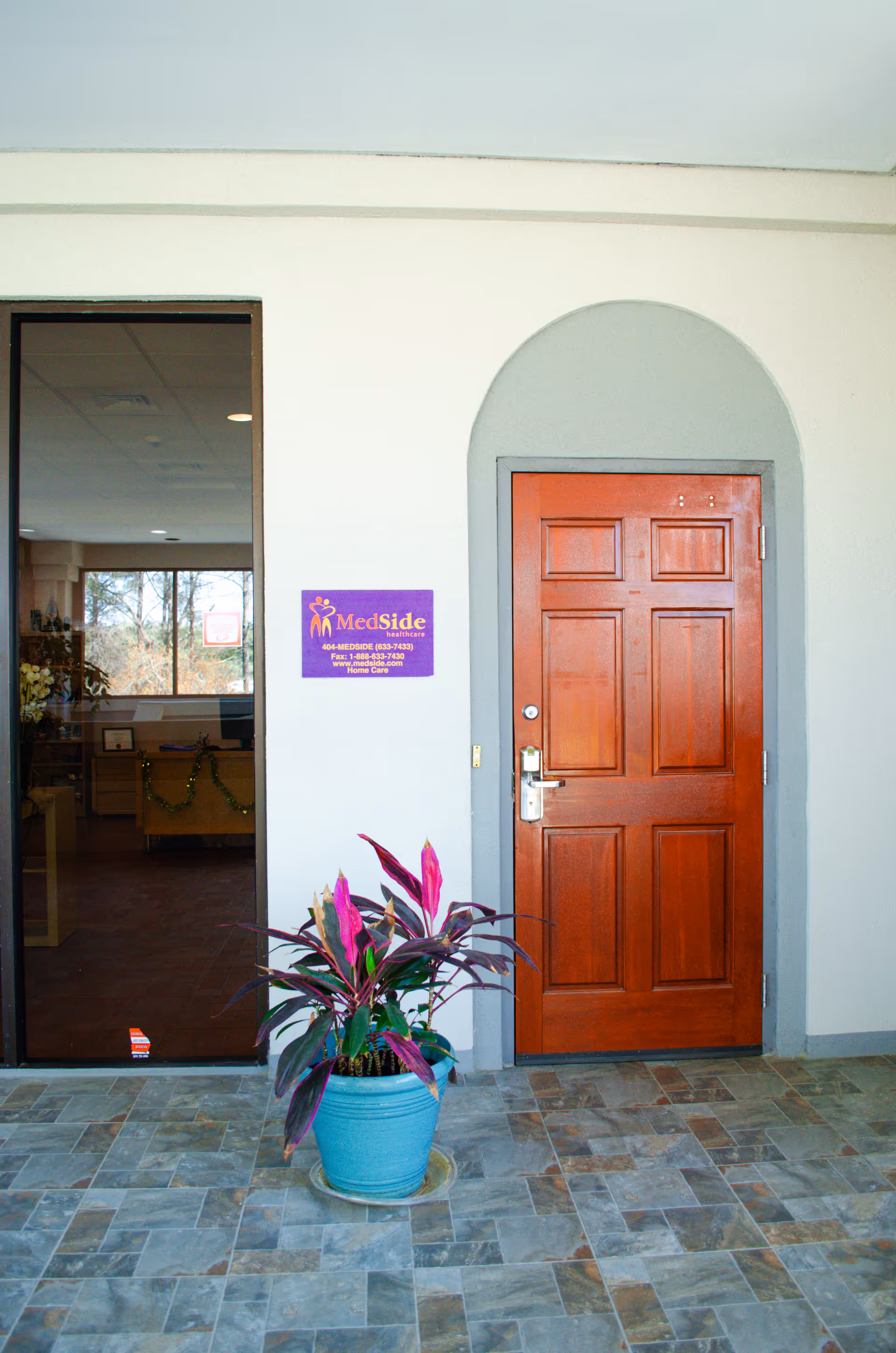 Entrance to MedSide Healthcare facility with wooden door, potted plant, and tile floor.