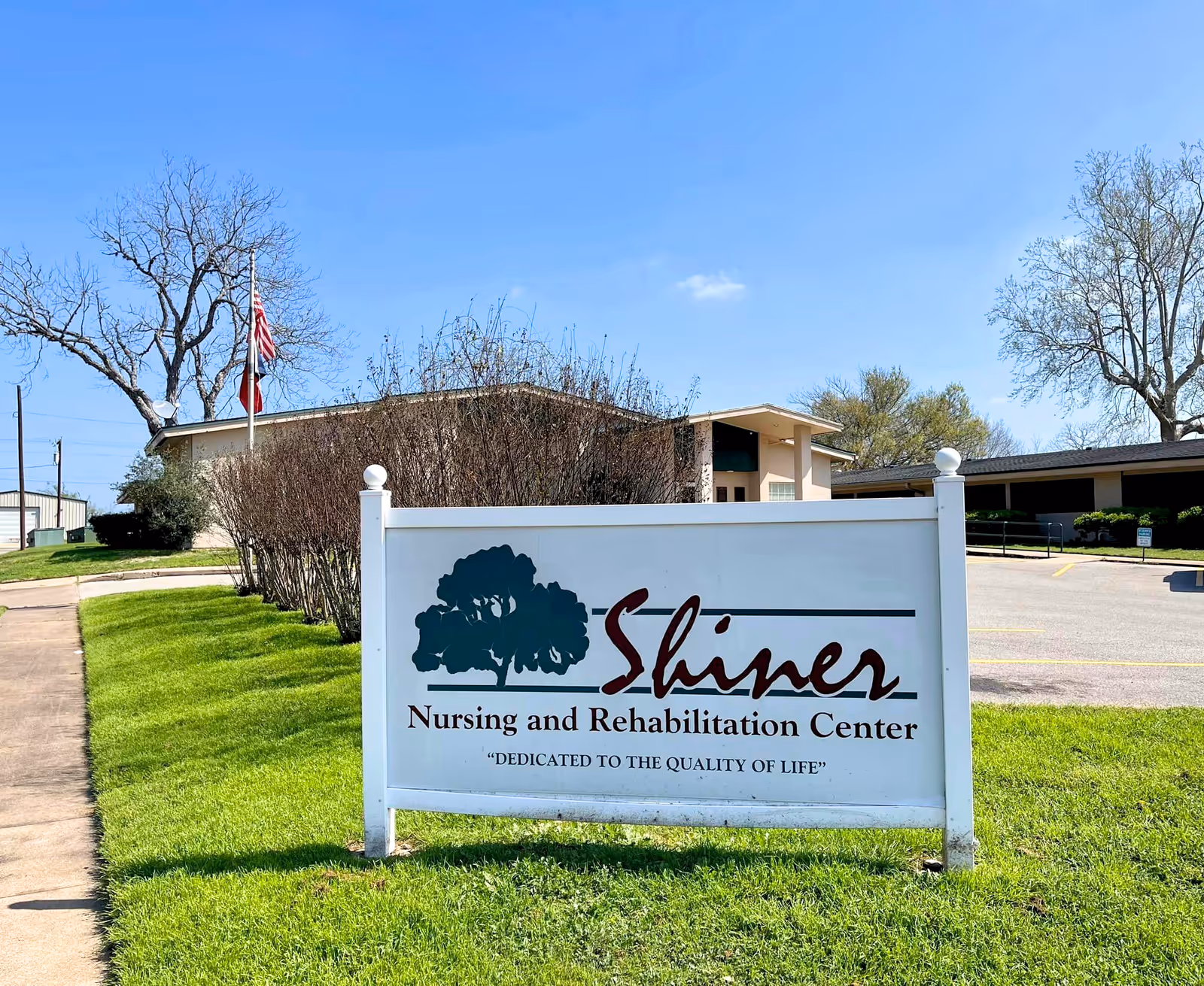 Outdoor view of Shiner Nursing and Rehabilitation Center sign with a building and parking lot in the background under a clear blue sky.