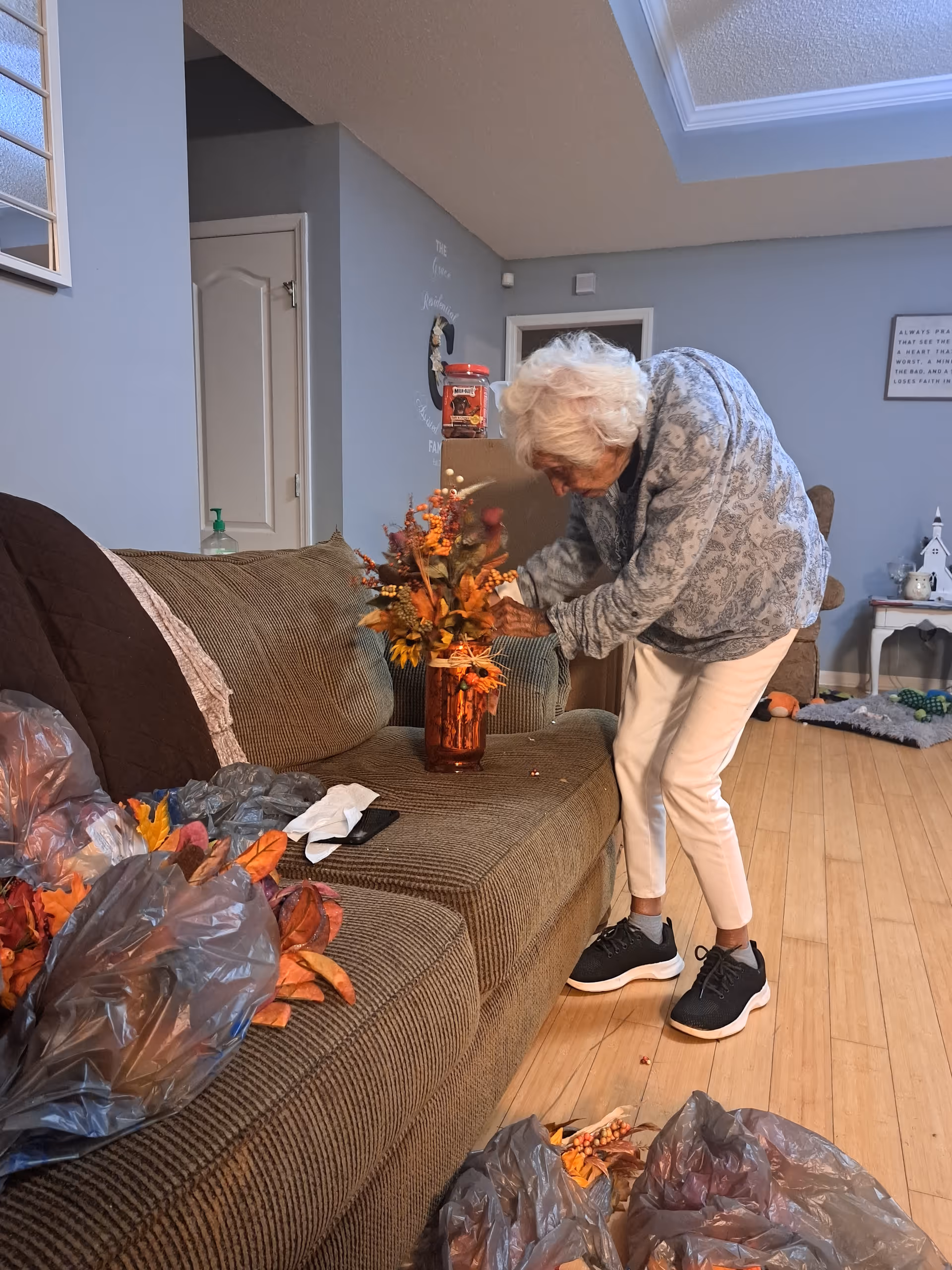 An elderly woman arranges orange flowers in a vase on a living room sofa.