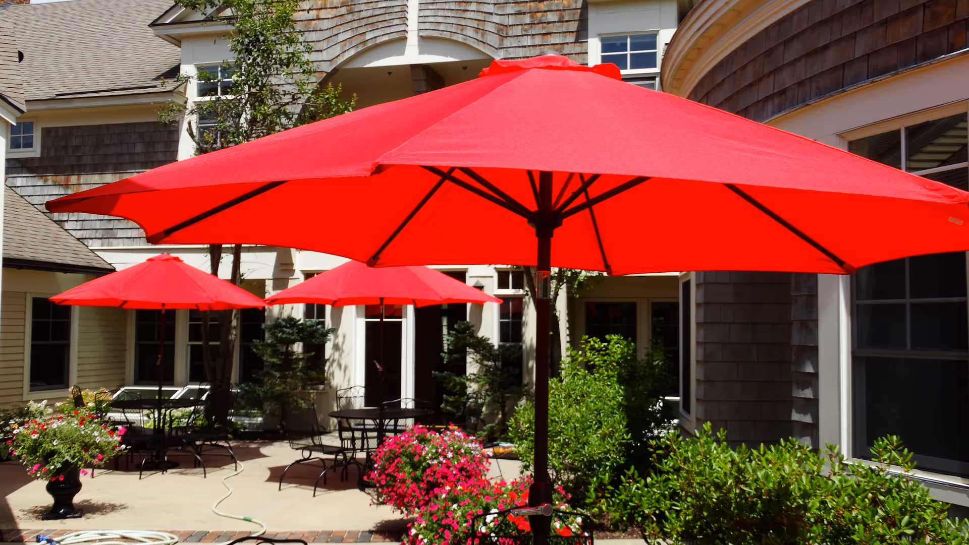 Outdoor patio area with several red umbrellas providing shade over black metal tables and chairs. The patio is surrounded by greenery and flowering plants, with a building featuring shingle siding and multiple windows in the background.