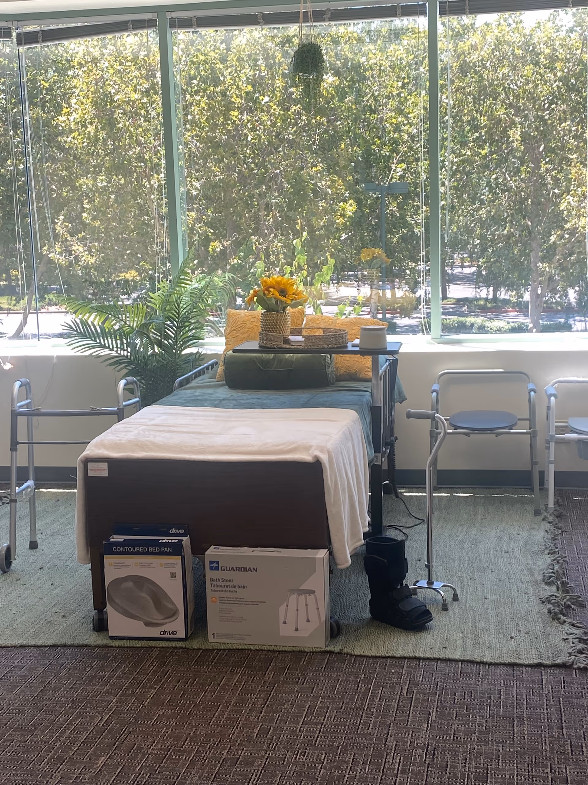 Care bed with supplies, a walker, and sunflowers in a bright windowed care room.