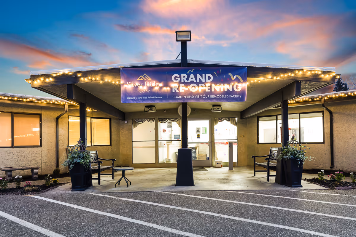 Exterior front entrance of Silver Ridge Care facility at dusk with a banner announcing grand reopening. The entrance is covered with a roof supported by pillars, decorated with string lights, benches, and potted plants on either side. The sky is colorful with clouds and the parking area is visible in front.