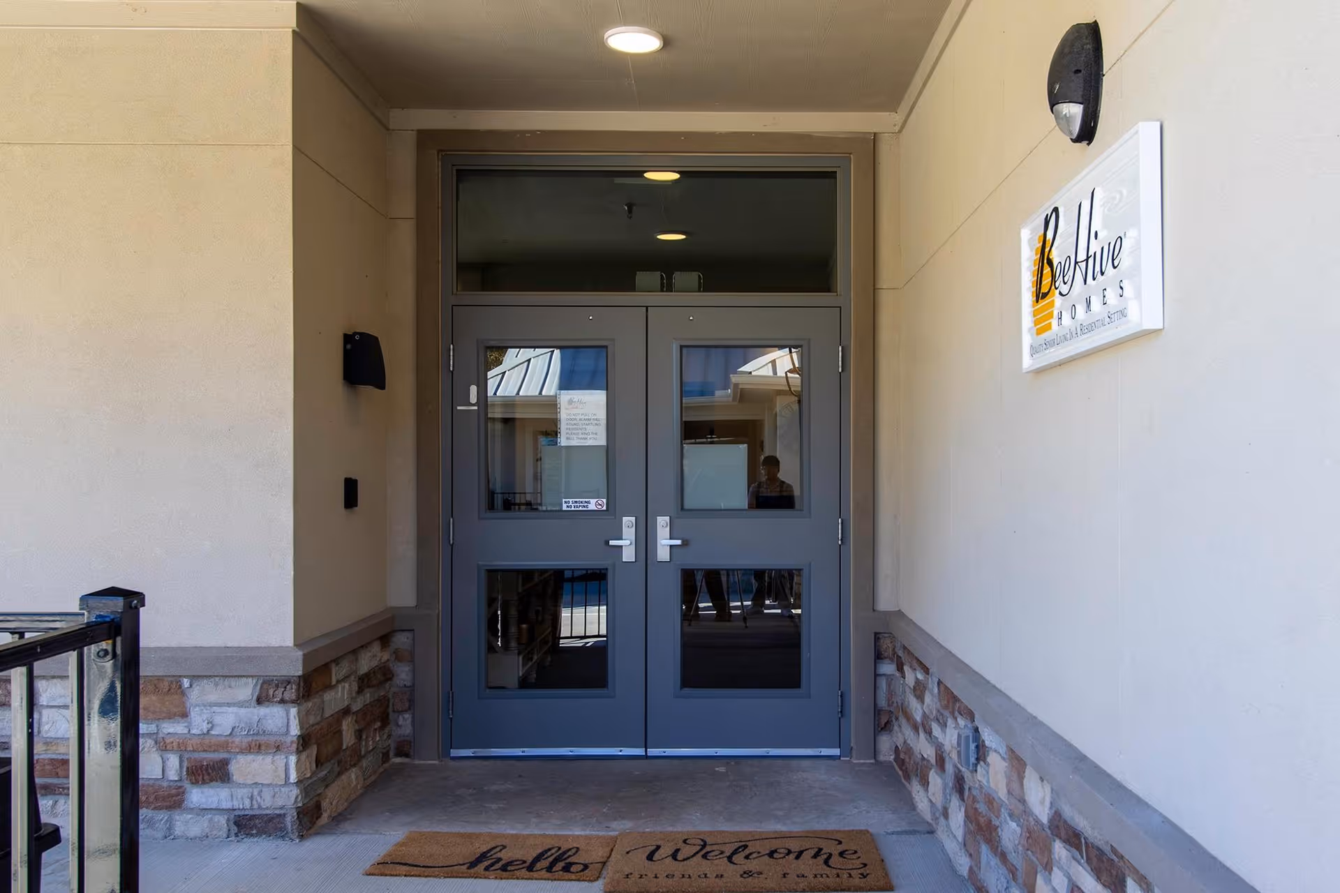 Entrance to BeeHive Homes showing double gray doors, a welcome mat, and a BeeHive Homes sign on the wall.