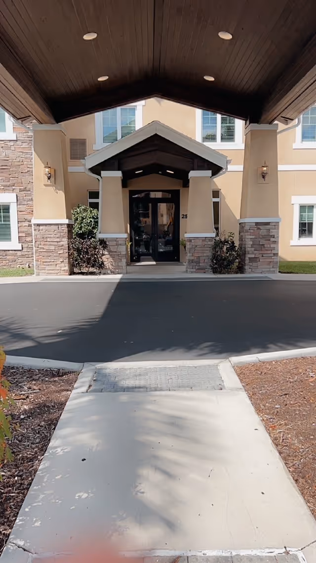 Entrance to a building with a covered driveway and a pathway leading up to double glass doors. The building exterior features beige walls with stone accents and several windows. There are small bushes on either side of the entrance.