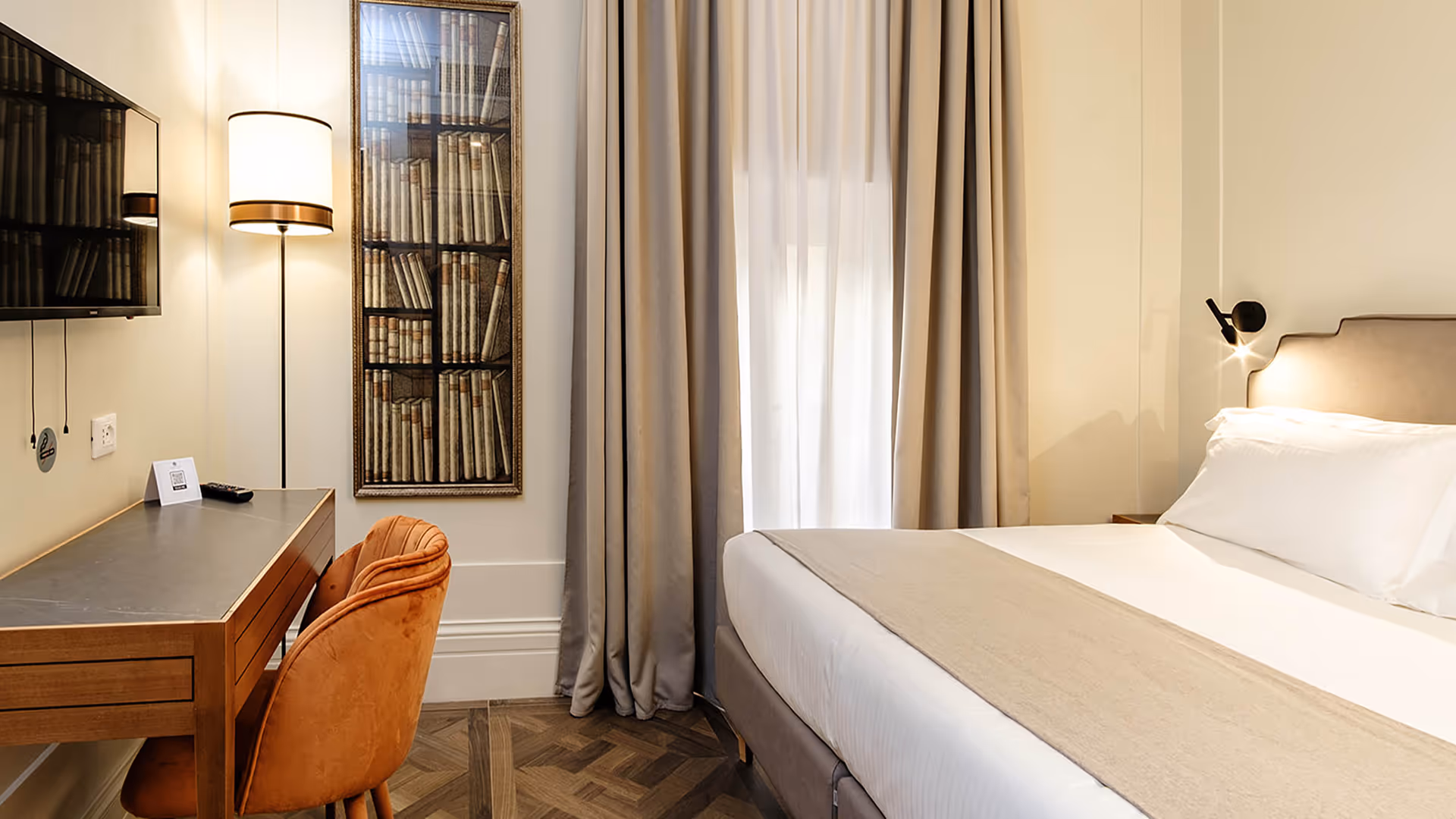 Well-lit hotel-style bedroom with a bed, desk, orange chair, and floor lamp.