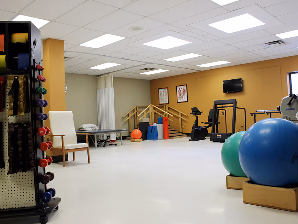 A physical therapy or rehabilitation room with exercise equipment including stationary bikes, a treadmill, large exercise balls, dumbbells, resistance bands, and a therapy table. The room has a white floor, beige and gray walls, fluorescent ceiling lights, and anatomical charts on the wall.