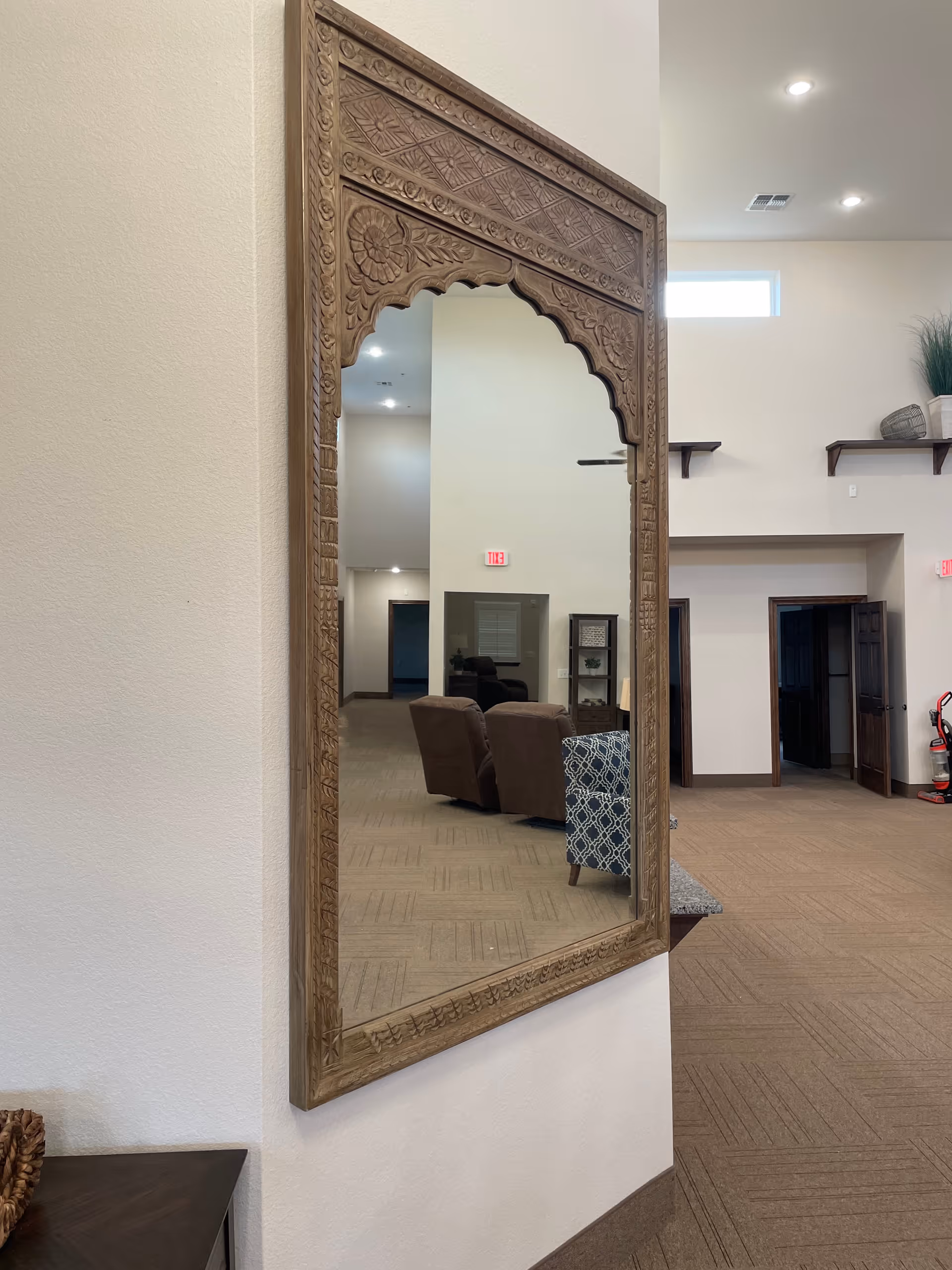 Ornate carved wooden mirror in a lounge hallway with chairs in background.