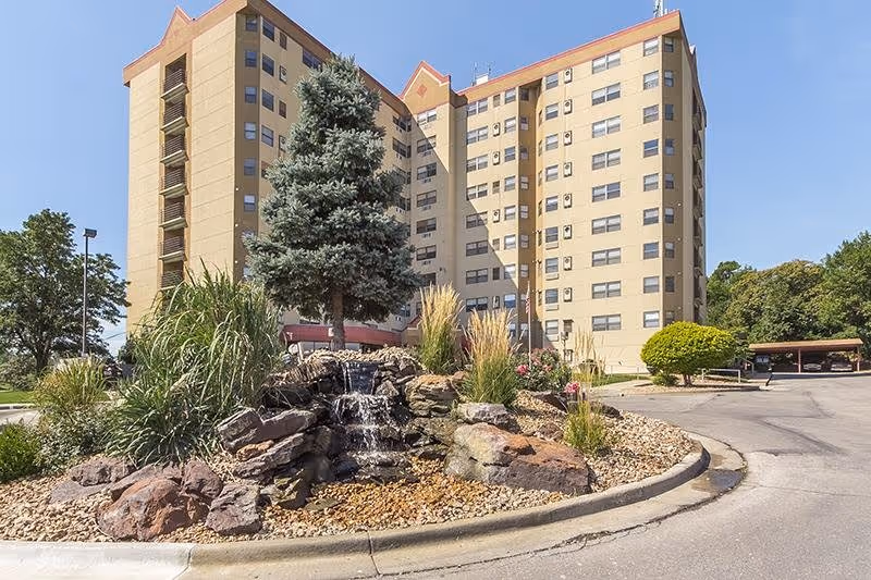 Tall beige senior living building with a landscaped rock fountain roundabout in front.
