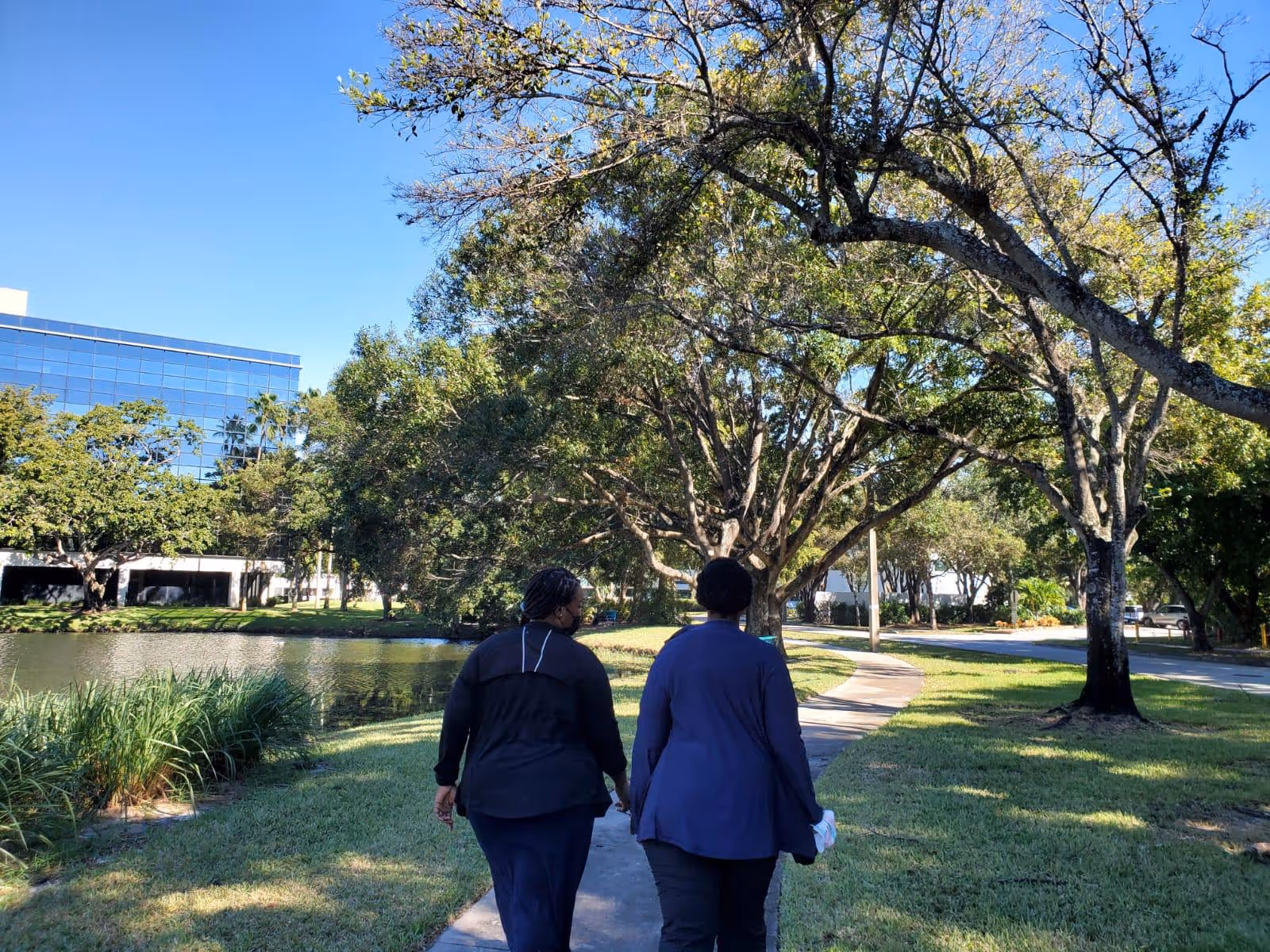 Two people walk along a sunny outdoor path beside a pond among trees.