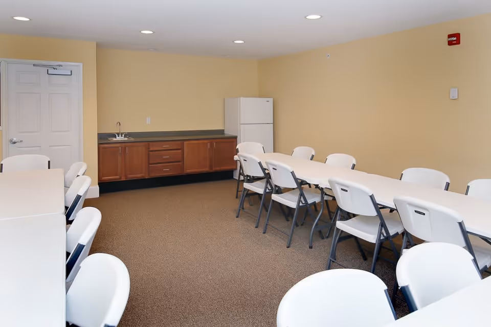A room with two long white tables arranged in an L shape, surrounded by white folding chairs. Against the far wall, there is a countertop with wooden cabinets below, a small sink, and a white refrigerator. The walls are painted light yellow and the floor is carpeted in brown.