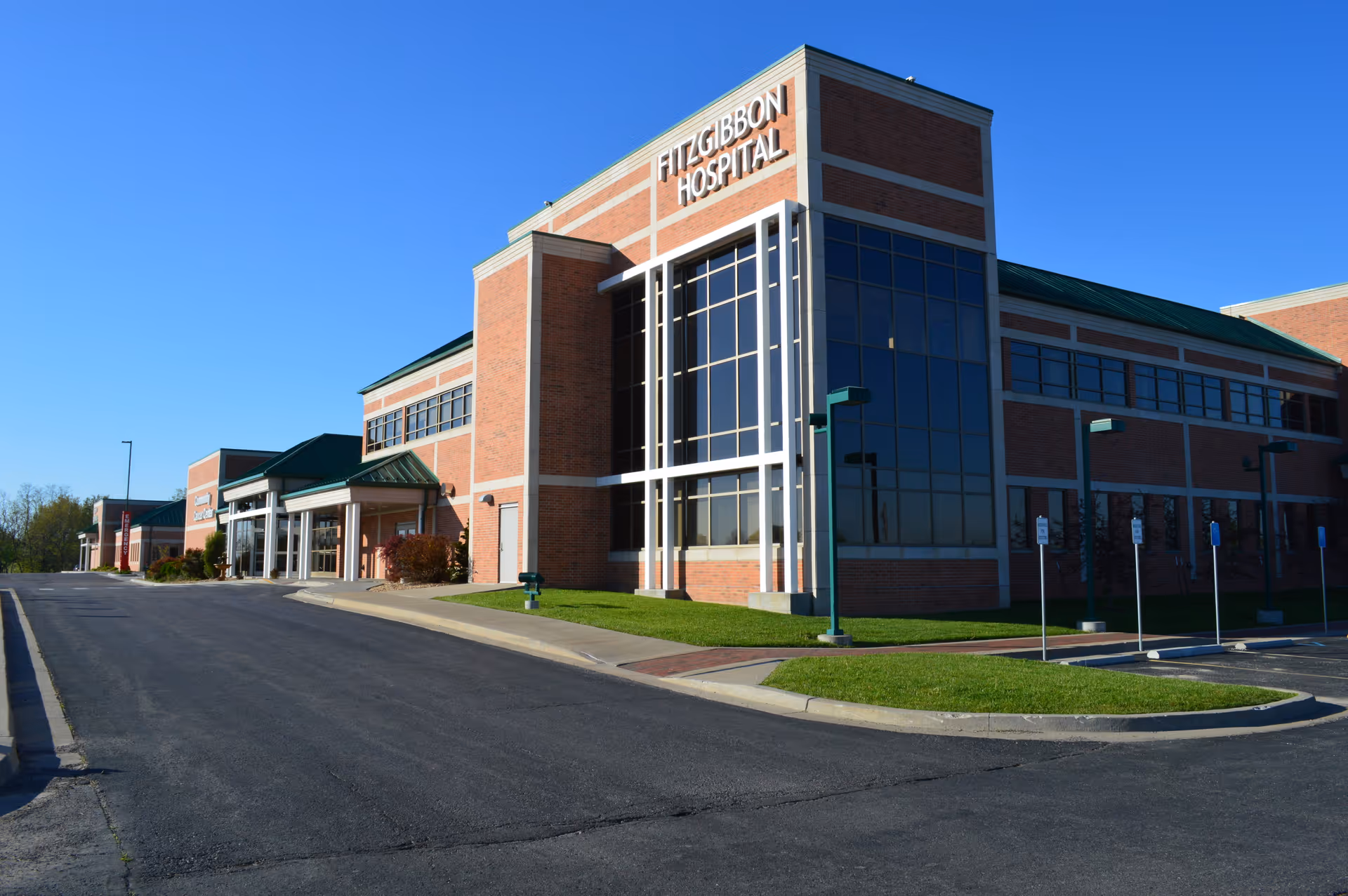 Front exterior of a brick hospital with a tall glass entrance and parking.