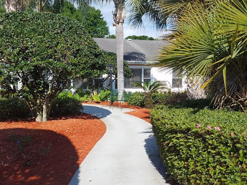 Curved concrete walkway through landscaped gardens with palm trees and shrubs leading to the front of a single-story building.