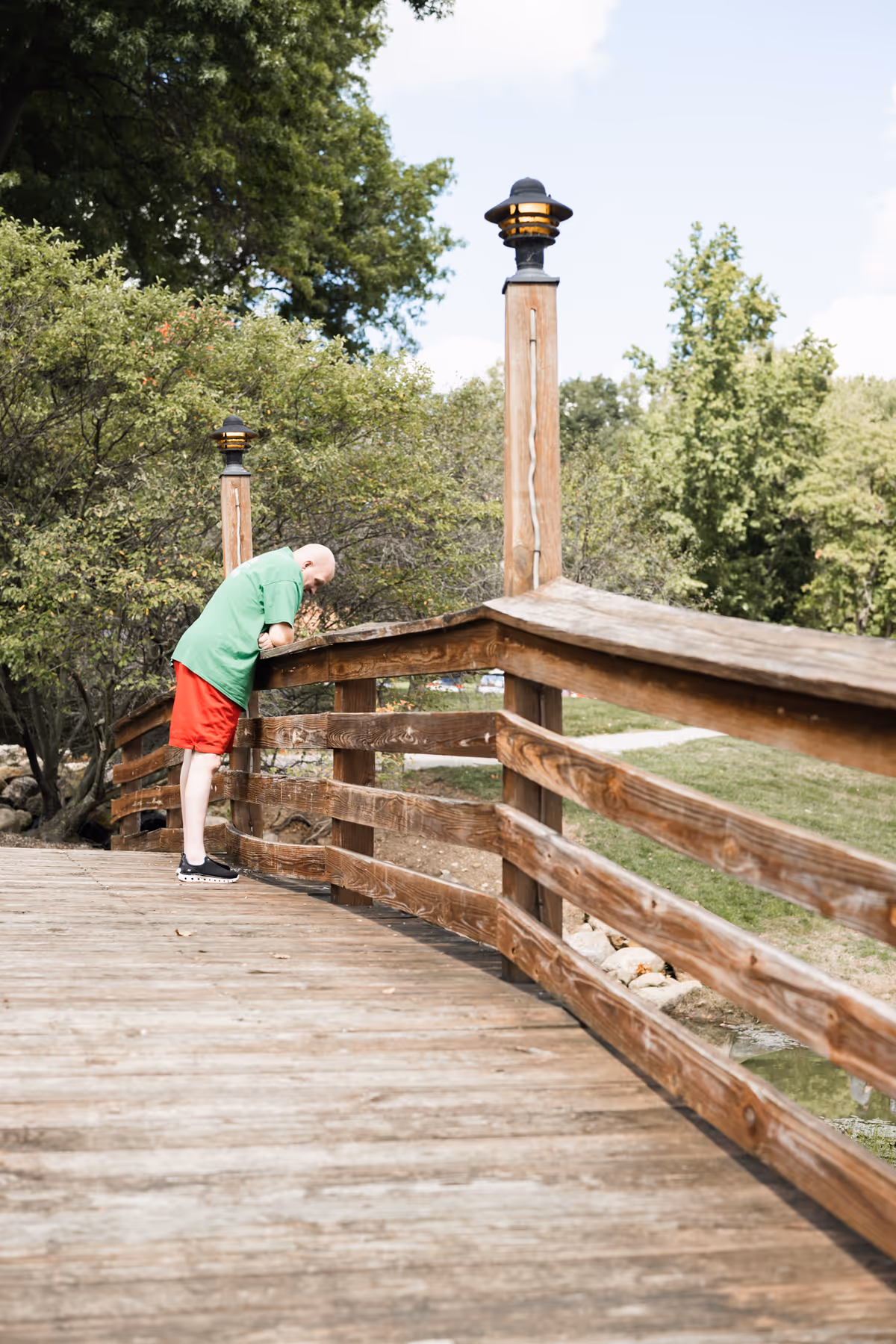 Person in green shirt and red shorts leans on a wooden railing on an outdoor bridge.