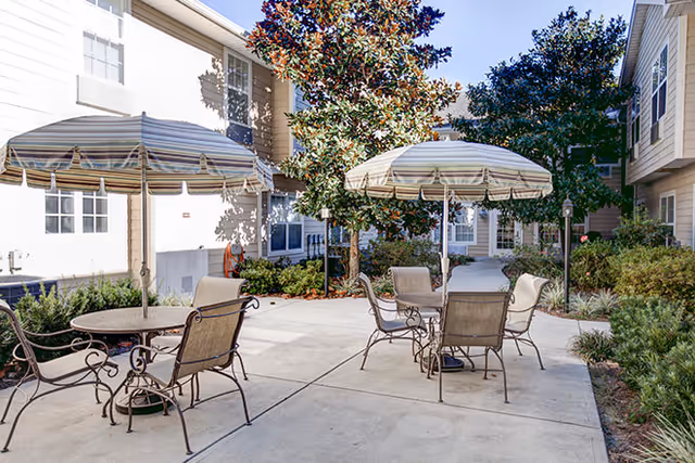 Outdoor courtyard area at The Island Cove at Hilton Head with two round tables, each shaded by striped umbrellas and surrounded by metal chairs. The courtyard is paved and bordered by shrubs and trees, with light-colored building walls in the background.