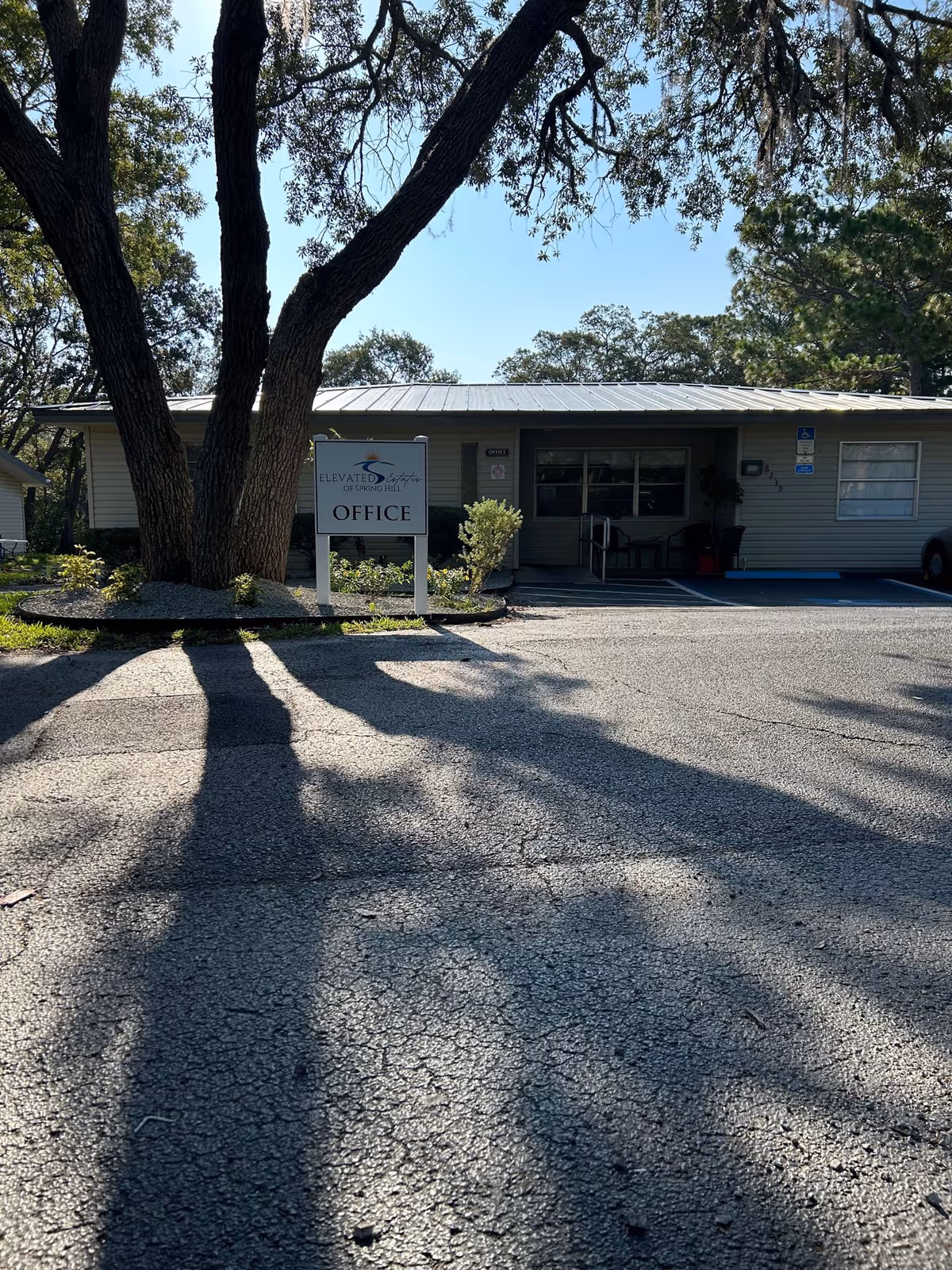 Exterior view of a single-story building with a sign in front that reads 'Elevated Estates of Spring Hill OFFICE'. The building has a metal roof and is surrounded by trees casting long shadows on the paved parking area in front.