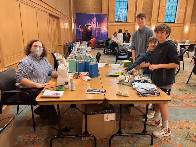 A group of people gathered around a long table in a large room with wood-paneled walls and tall windows. One person wearing a mask is seated at the table with a sewing machine, while three boys stand on the opposite side of the table with various craft materials and bags. Other people are visible in the background, and the room has a patterned carpet.