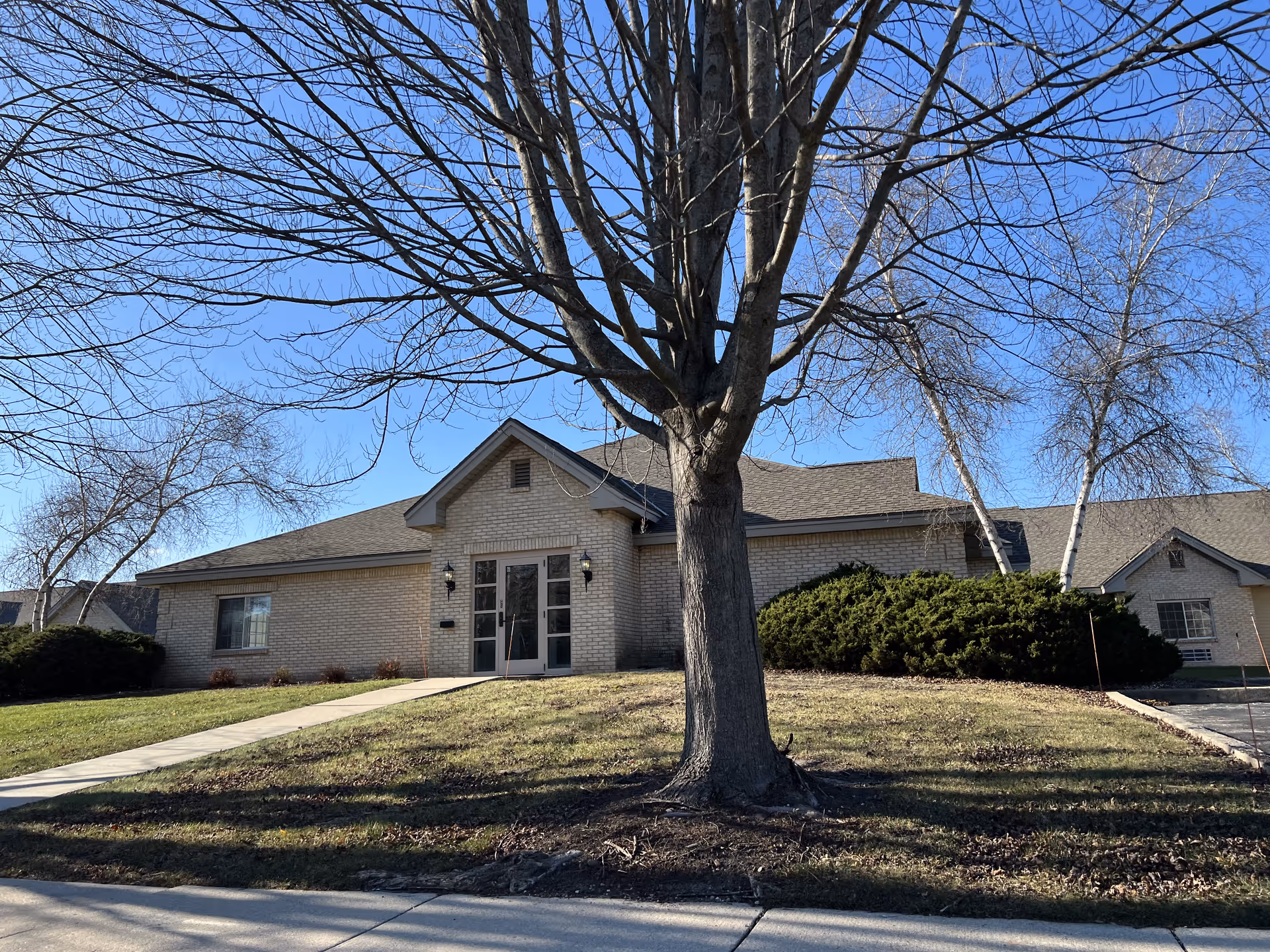 A single-story brick building with a peaked roof and double glass doors at the entrance. In front of the building is a large leafless tree and a grassy lawn with a concrete walkway leading to the entrance. The sky is clear and blue.