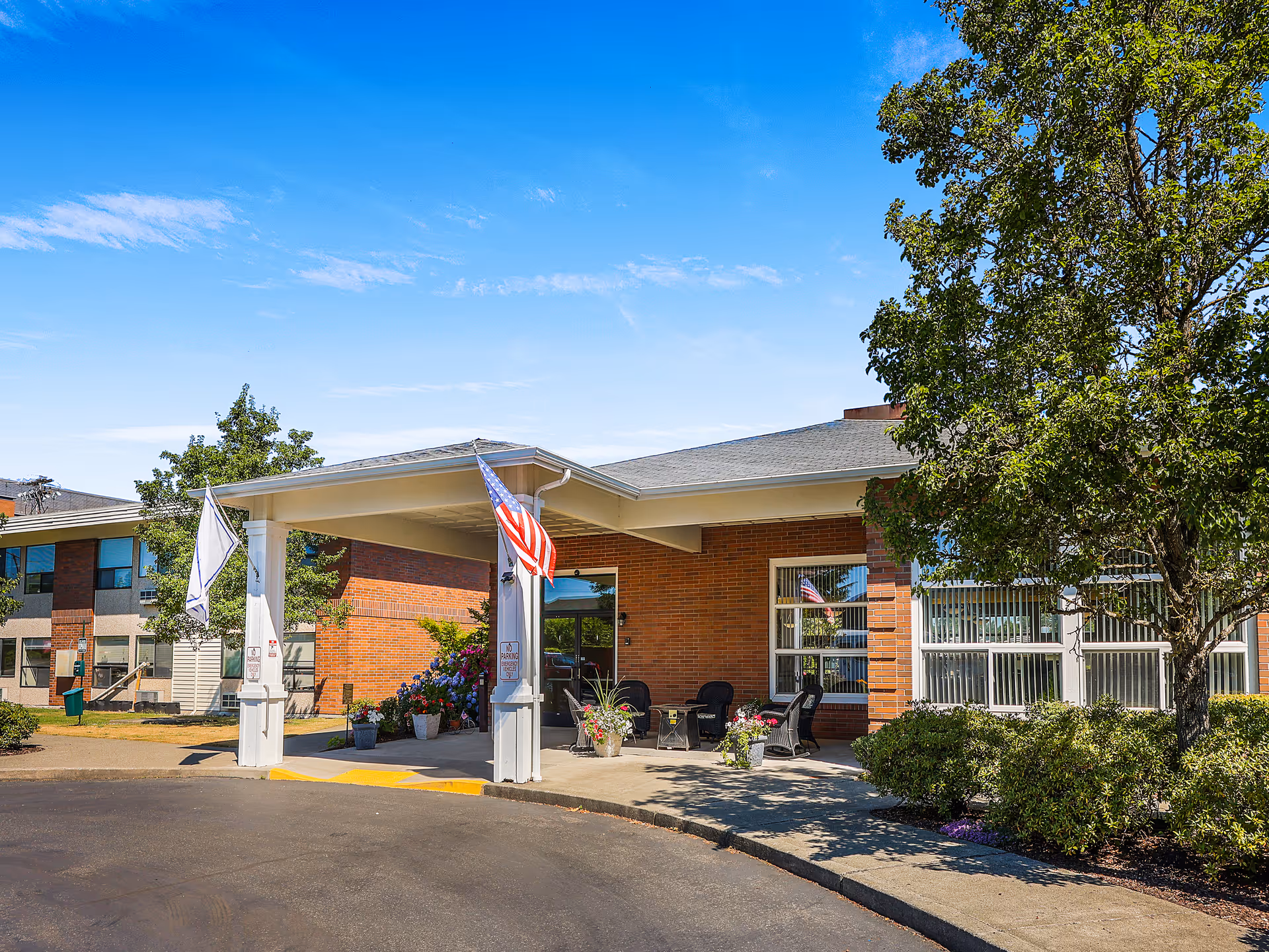 Front entrance of a brick assisted living facility with a covered porte-cochère, an American flag, seating and landscaping.