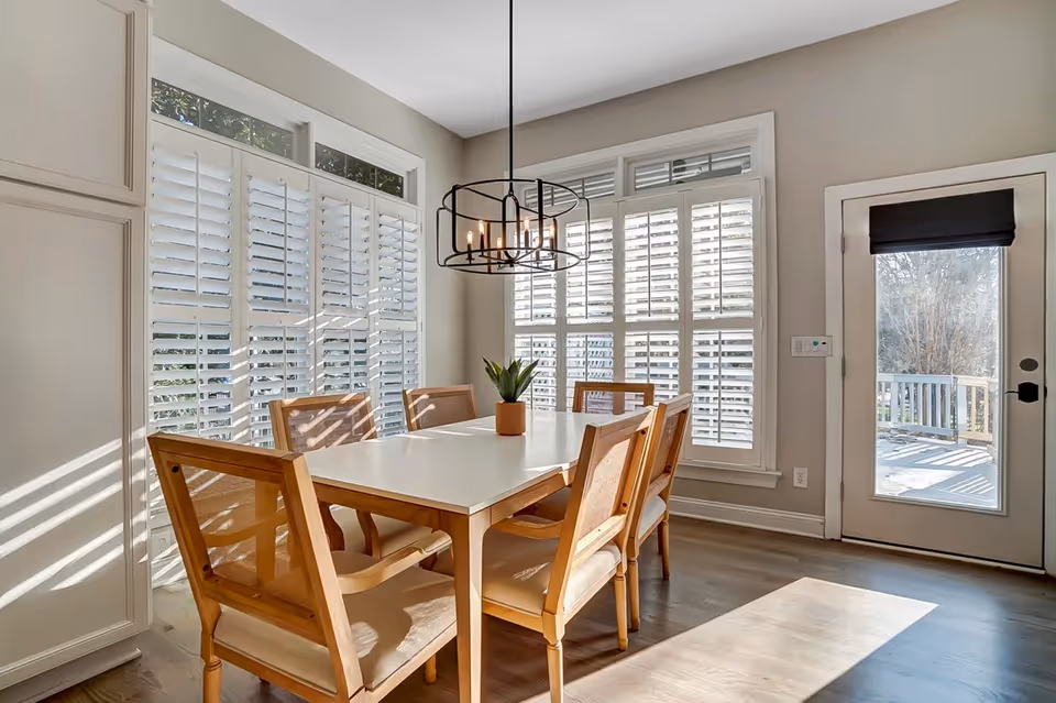 Bright dining room with white shutters, a white table, six wooden chairs, and a glass door.