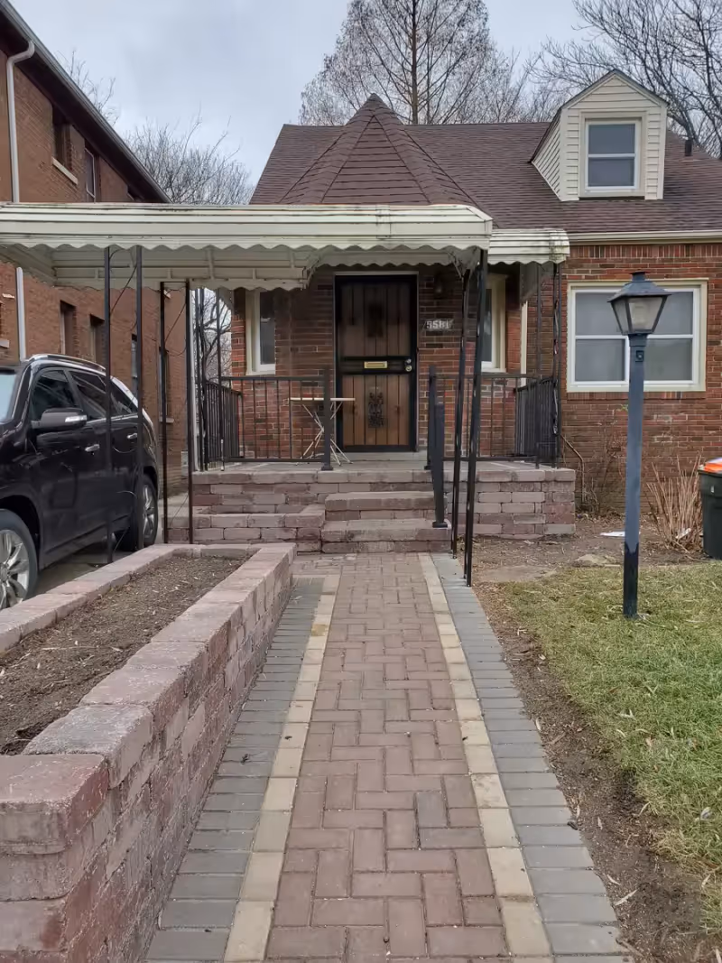 Paved walkway leading to the covered front porch of a brick house with steps, a lamp post on the right and a parked car on the left.