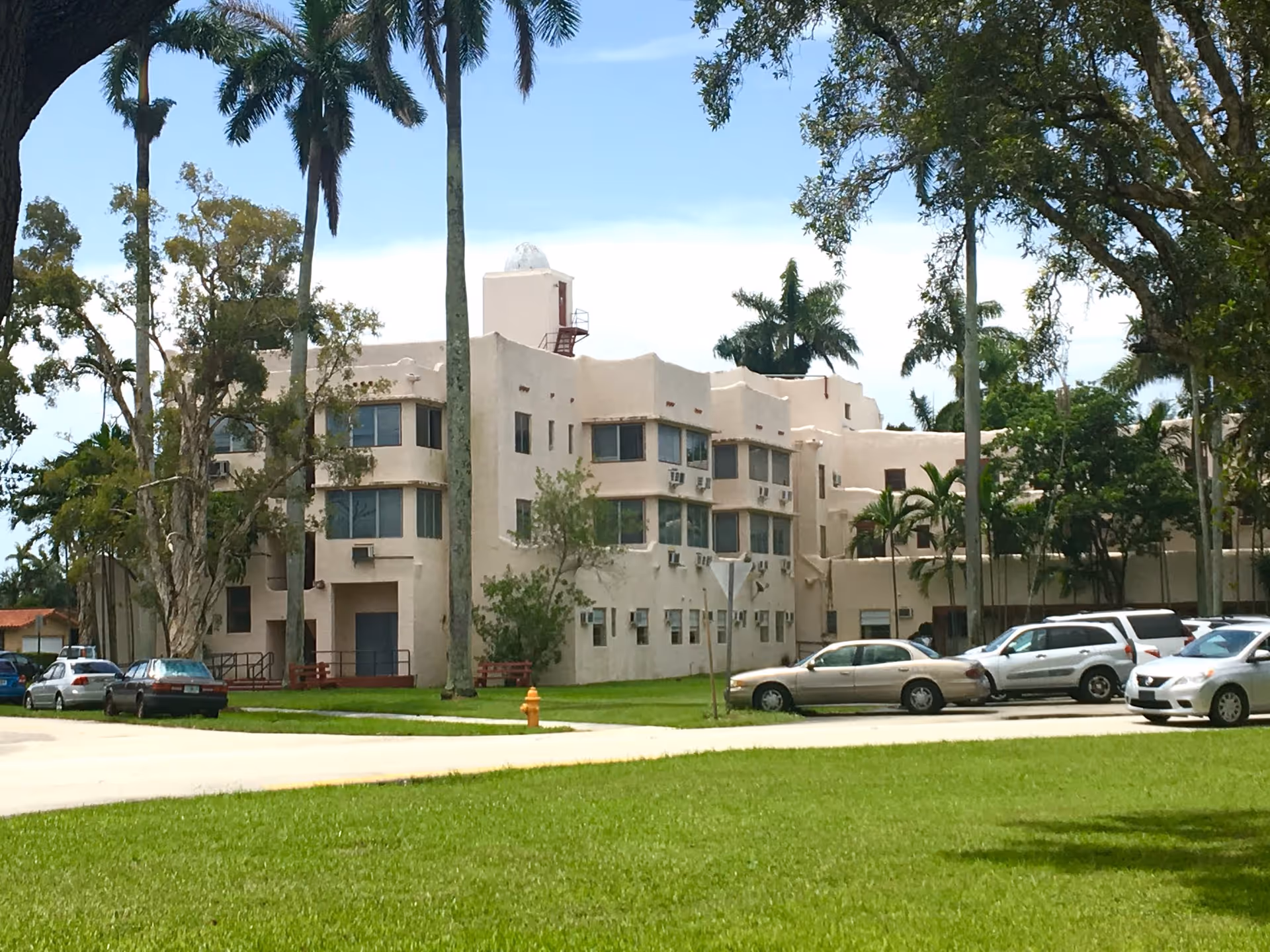 Exterior view of a beige senior living center with palm trees and parked cars.
