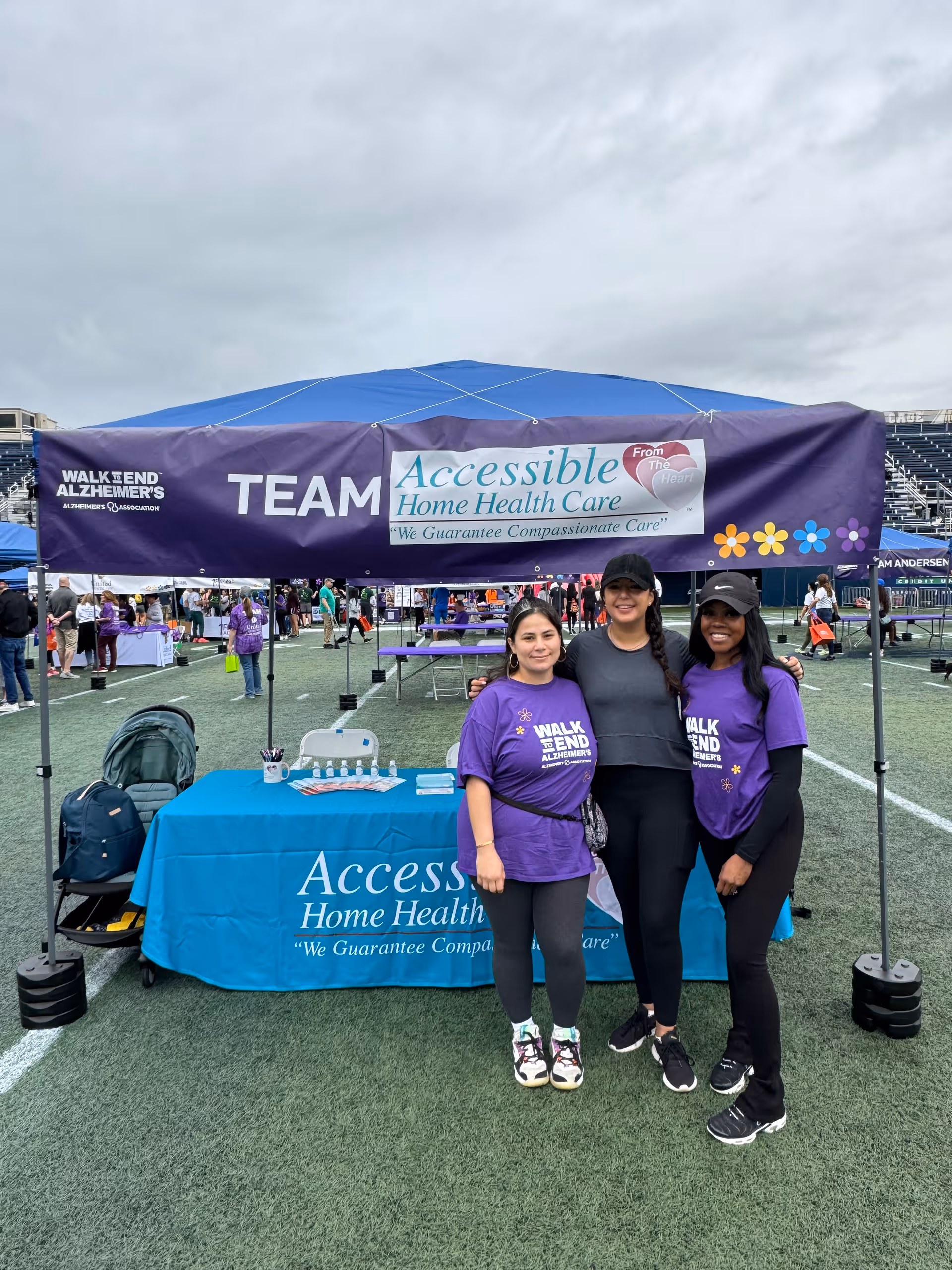 Three women in purple shirts pose beside an Accessible Home Health Care tent outdoors.