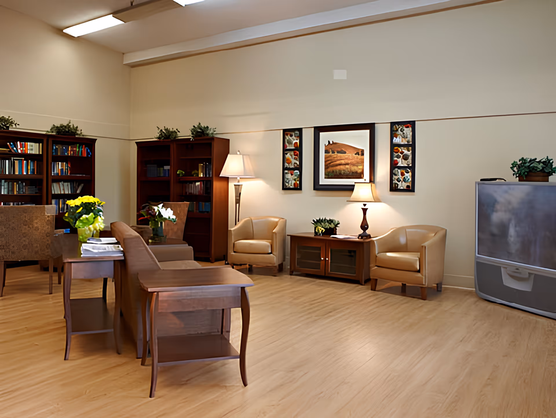Bright senior living common room with seating, bookshelves, lamps, artwork, and a large TV.