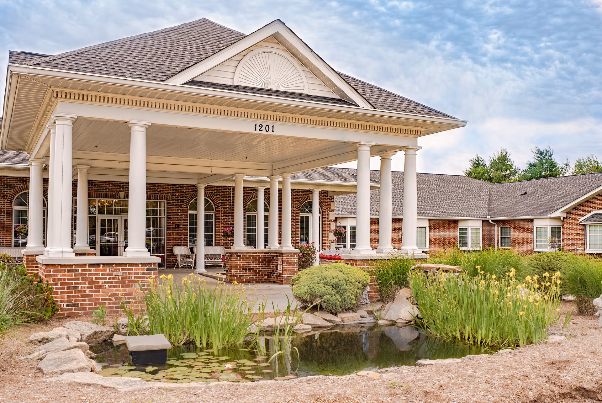 Senior living facility exterior with brick walls, white columns, front entrance, and a landscaped pond.