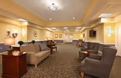 A spacious and well-lit living room area in an assisted living facility featuring multiple seating arrangements including sofas and armchairs, side tables with lamps, framed artwork on the walls, and a carpeted floor with a patterned design.