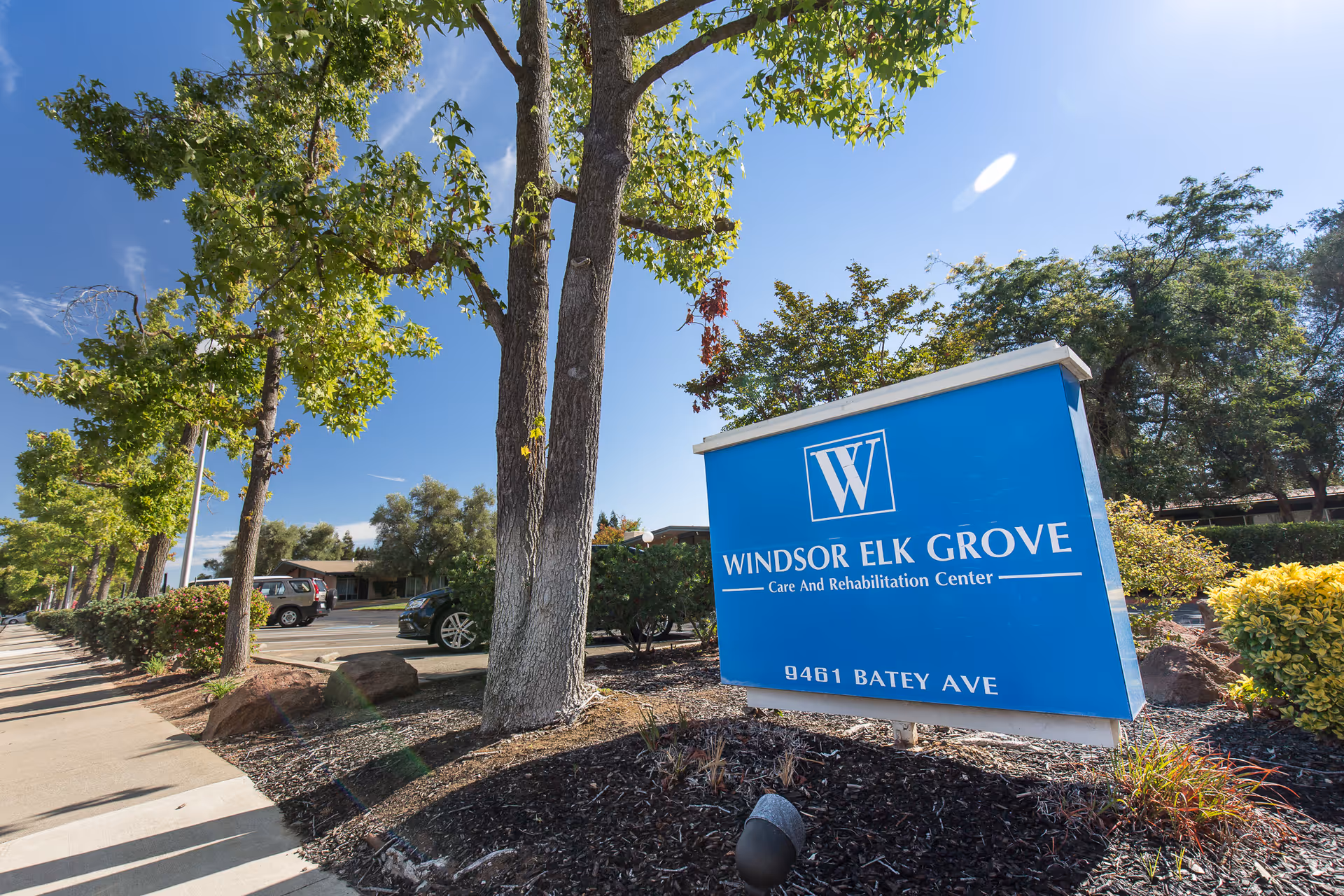 Outdoor view of Windsor Elk Grove Care and Rehabilitation Center sign with trees, bushes, parked cars, and a clear blue sky in the background.