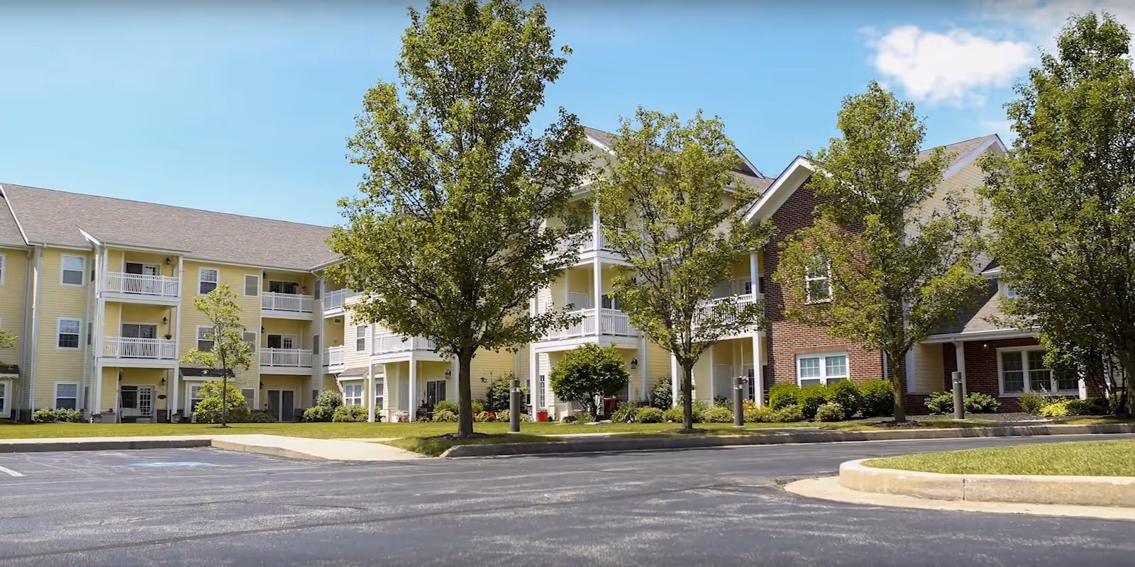 Exterior view of a multi-story retirement community building with yellow siding and brick accents, surrounded by green trees and landscaping under a clear blue sky.