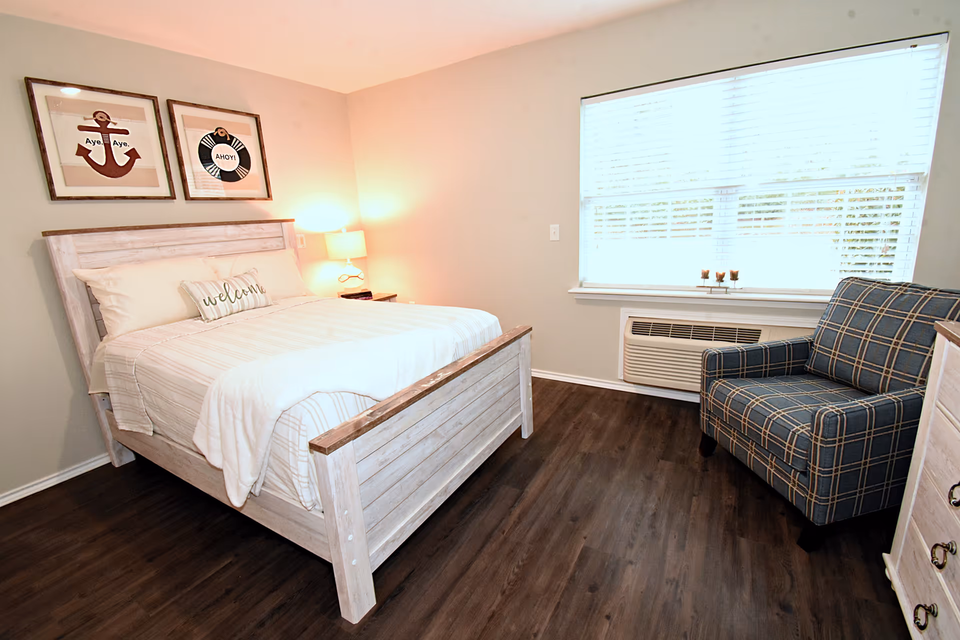 A bright and cozy bedroom with a wooden bed frame, white bedding, and a decorative pillow that says 'welcome'. There are two framed nautical-themed pictures on the wall above the bed. A nightstand with a lamp is next to the bed. A plaid upholstered armchair is positioned near a large window with white blinds. The room has dark wood flooring and light-colored walls.