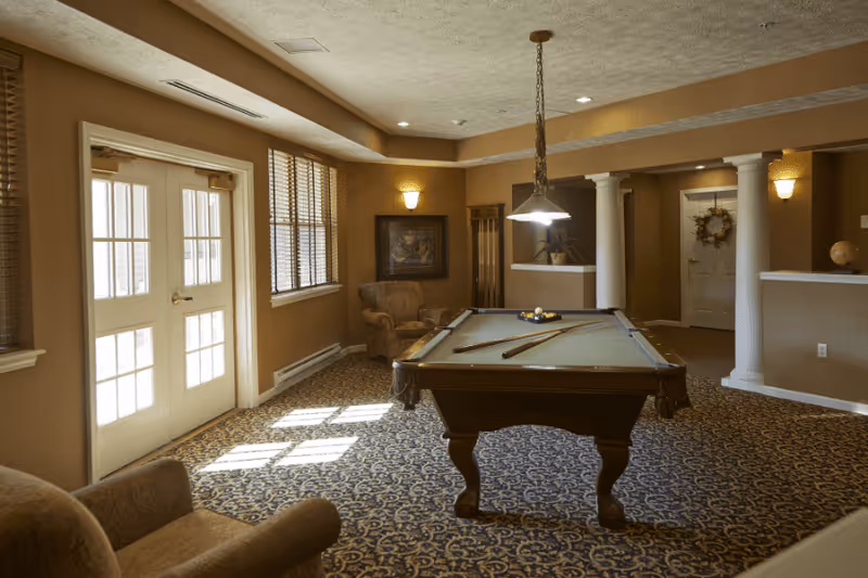A cozy indoor recreation room with a pool table in the center, two pool cues and a rack of balls on the table, beige armchairs, patterned carpet, large windows with blinds letting in natural light, decorative columns, and warm wall sconces.