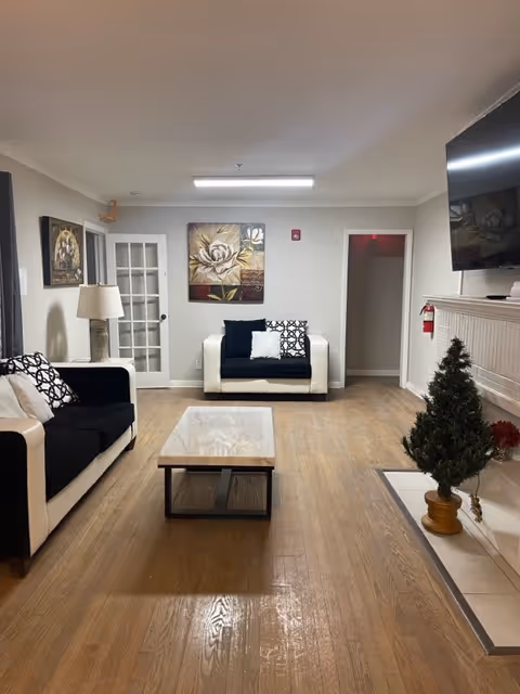 Bright senior living lounge with white sofas, coffee table, artwork, and a fireplace mantel.