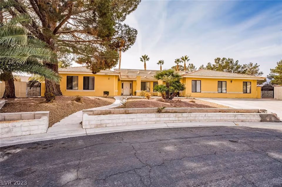 Front exterior view of a single-story yellow building with a tiled roof, surrounded by desert landscaping including palm trees and other shrubs, under a partly cloudy sky.