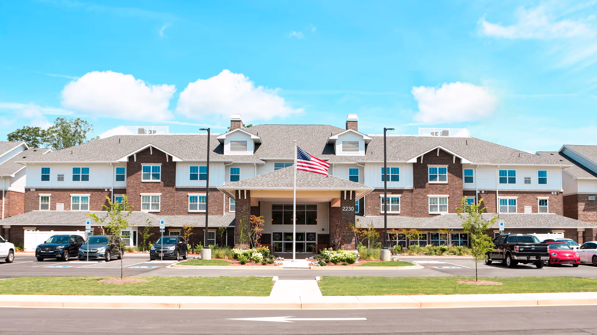 Front view of a brick senior living building with entrance, flag, and parked cars.