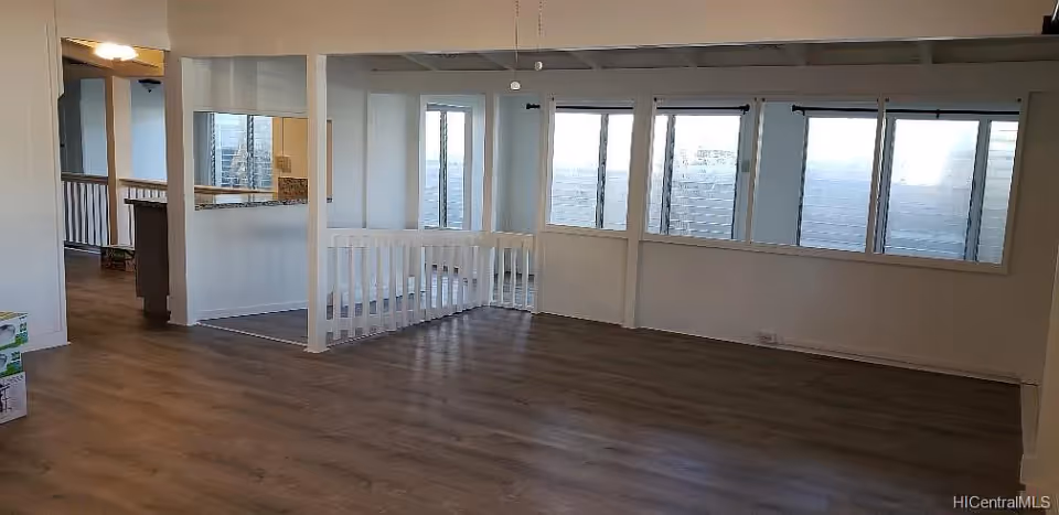 An empty interior room with wooden flooring and white walls. The room features a railing around a stairwell or open area in the center, multiple windows along one wall, and an open doorway leading to another space. The area is well-lit with natural light coming through the windows.