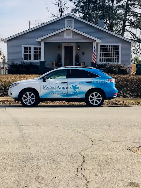 Front view of a gray, single-story home with a blue Visiting Angels car parked.