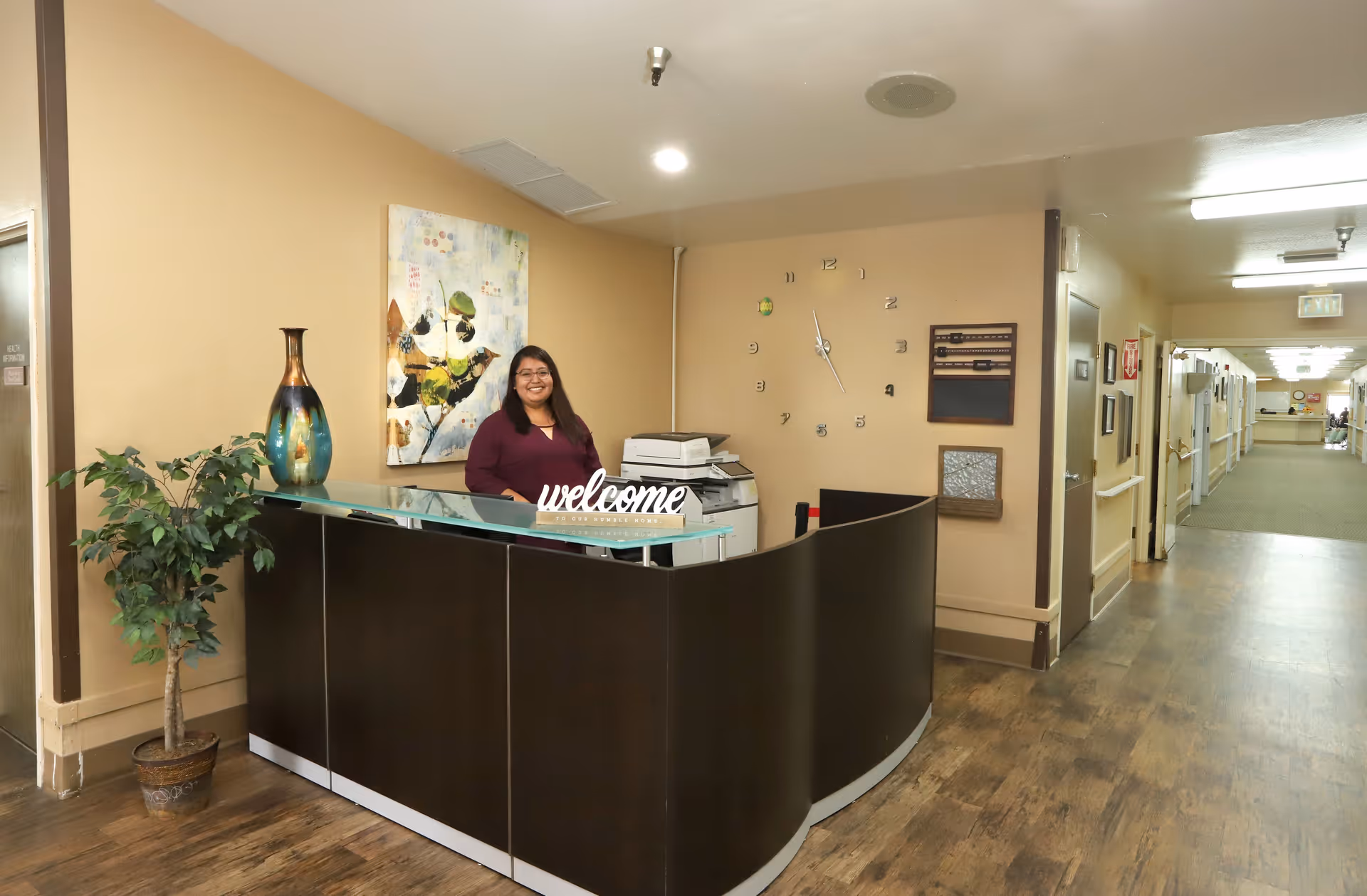 Reception area inside a senior living facility with a smiling woman standing behind a curved black desk that has a 'welcome' sign on it. The area features a potted plant, a decorative vase, a wall clock with modern numbers, and a hallway extending to the right with doors and handrails along the walls.