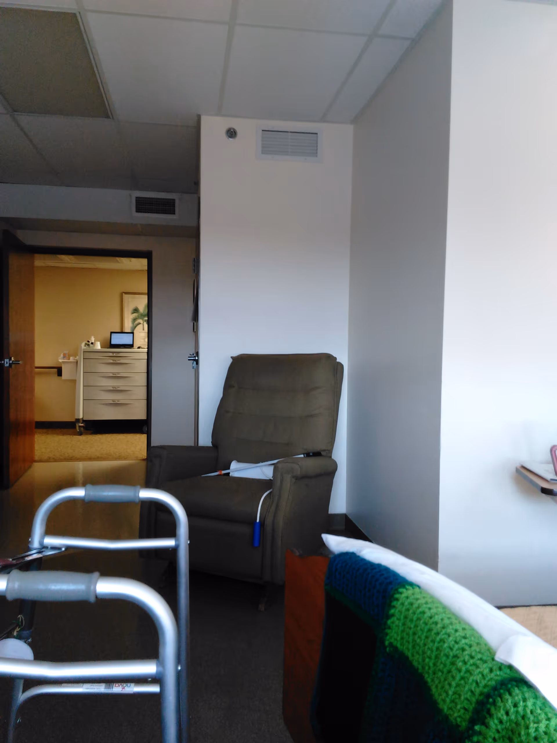 Gray recliner, walker, and colorful blanket in a care facility interior with an open doorway.