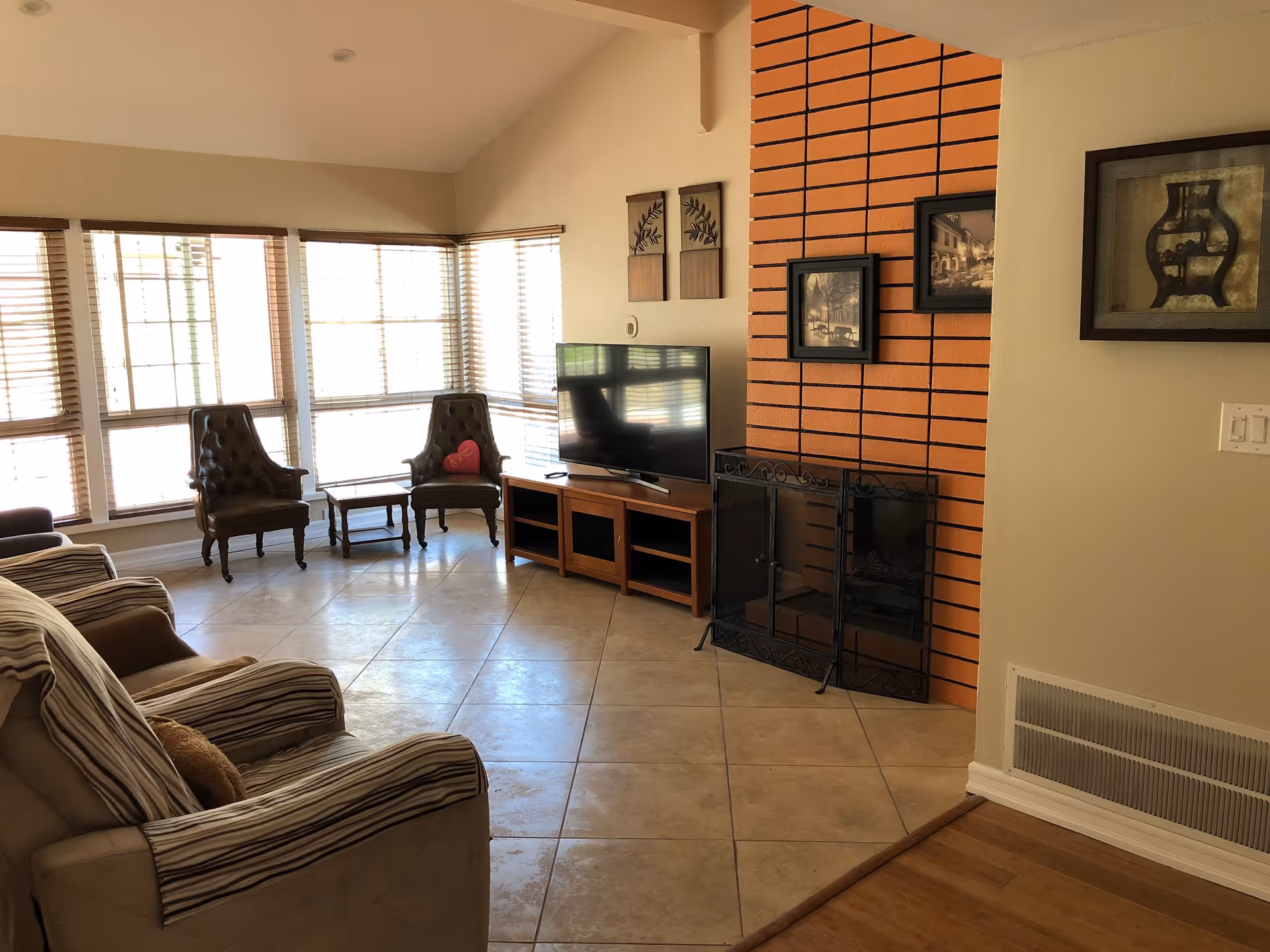 Sunlit living room with large bay windows, upholstered chairs, a TV on a stand, and a brick fireplace.