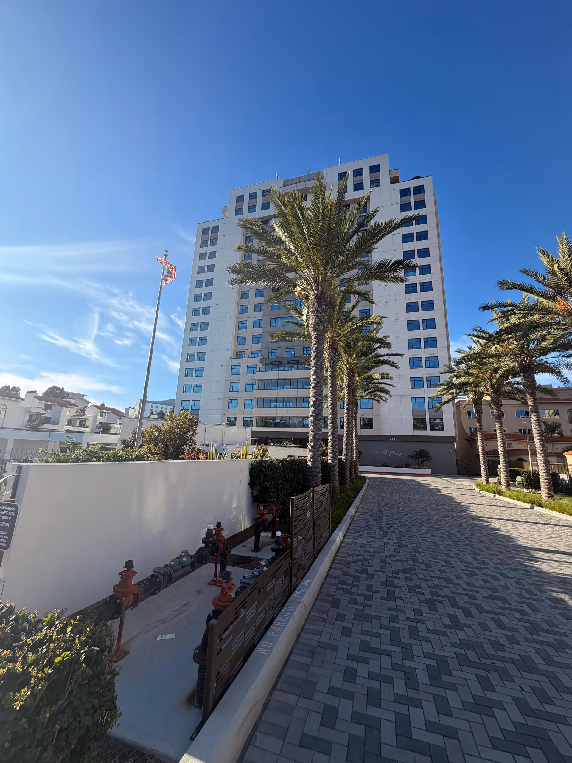 Tall white senior living building with palm trees along a paved entry path.