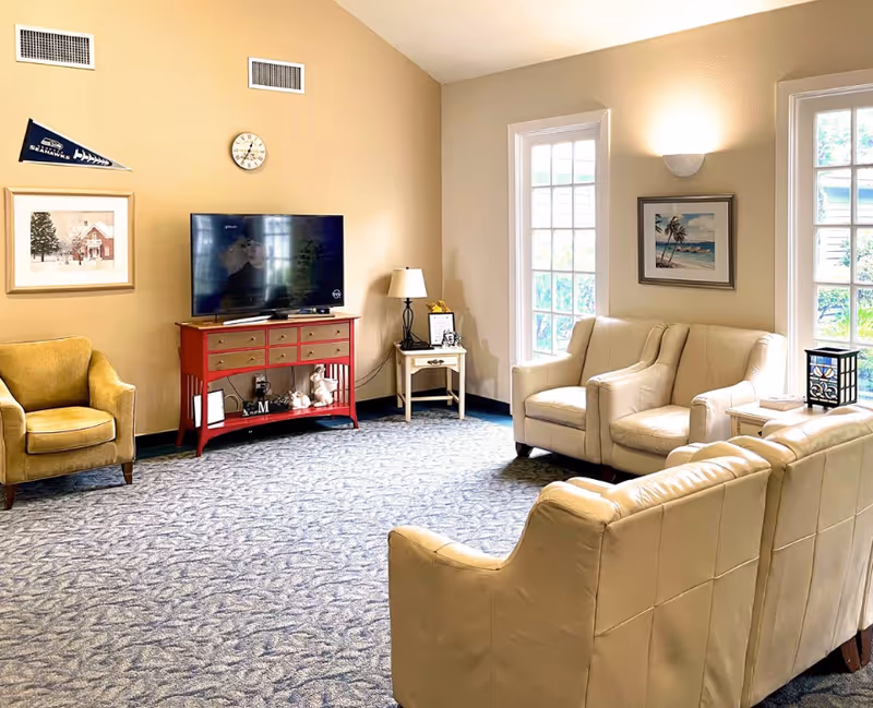 Cozy senior living lounge with cream seating, a red TV console, and large windows.