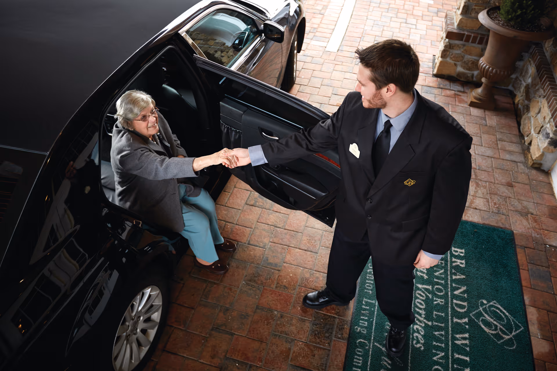 A valet helps an elderly woman step out of a black car at the entrance of a senior living facility.