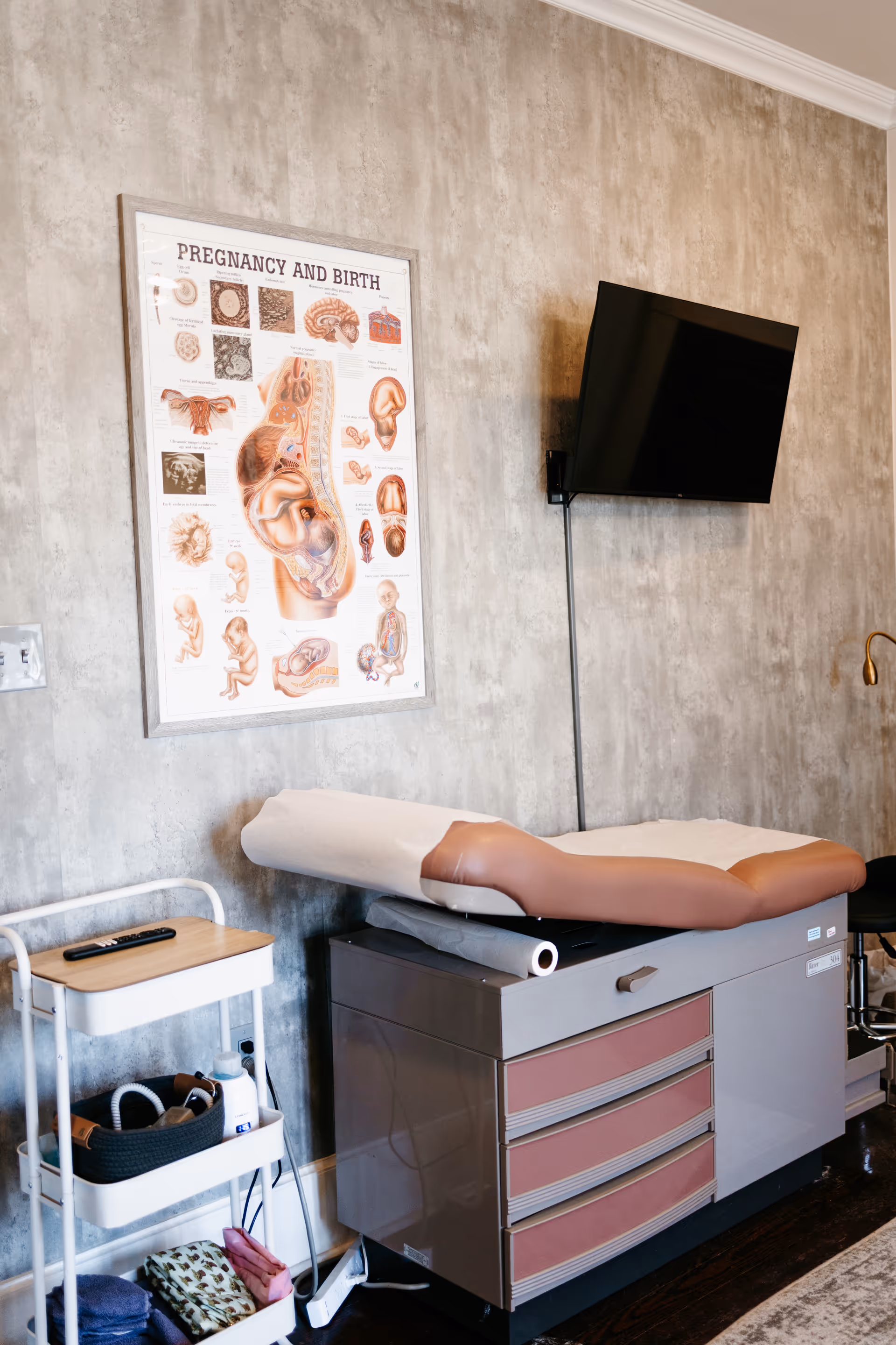 Interior exam room with a medical mannequin leg on a cabinet and wall-mounted TV.