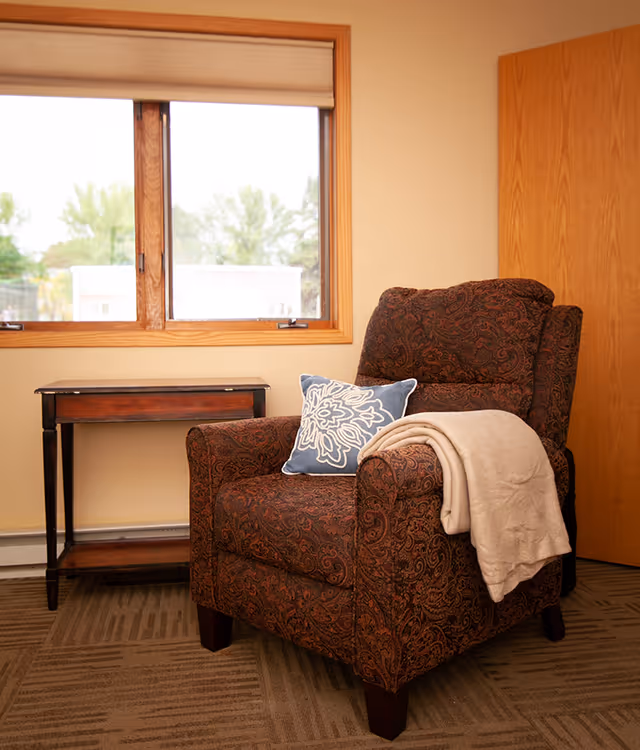 Cozy patterned armchair with blue pillow and beige blanket by the window.