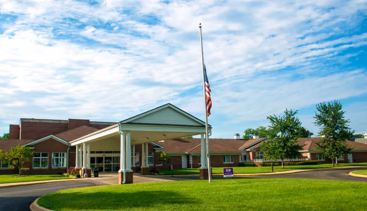 Exterior view of a senior living facility with a covered entrance, lawn, and flagpole.
