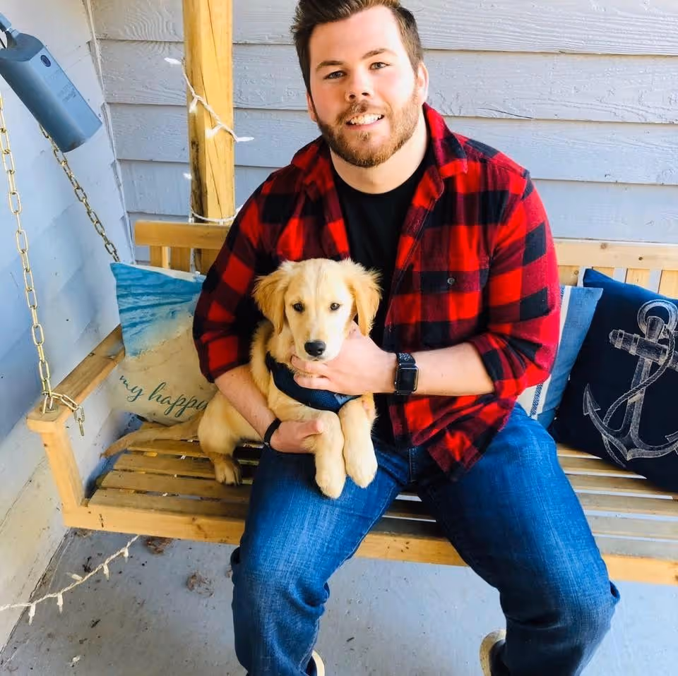 Man in red plaid shirt sits on a wooden porch swing with a puppy.